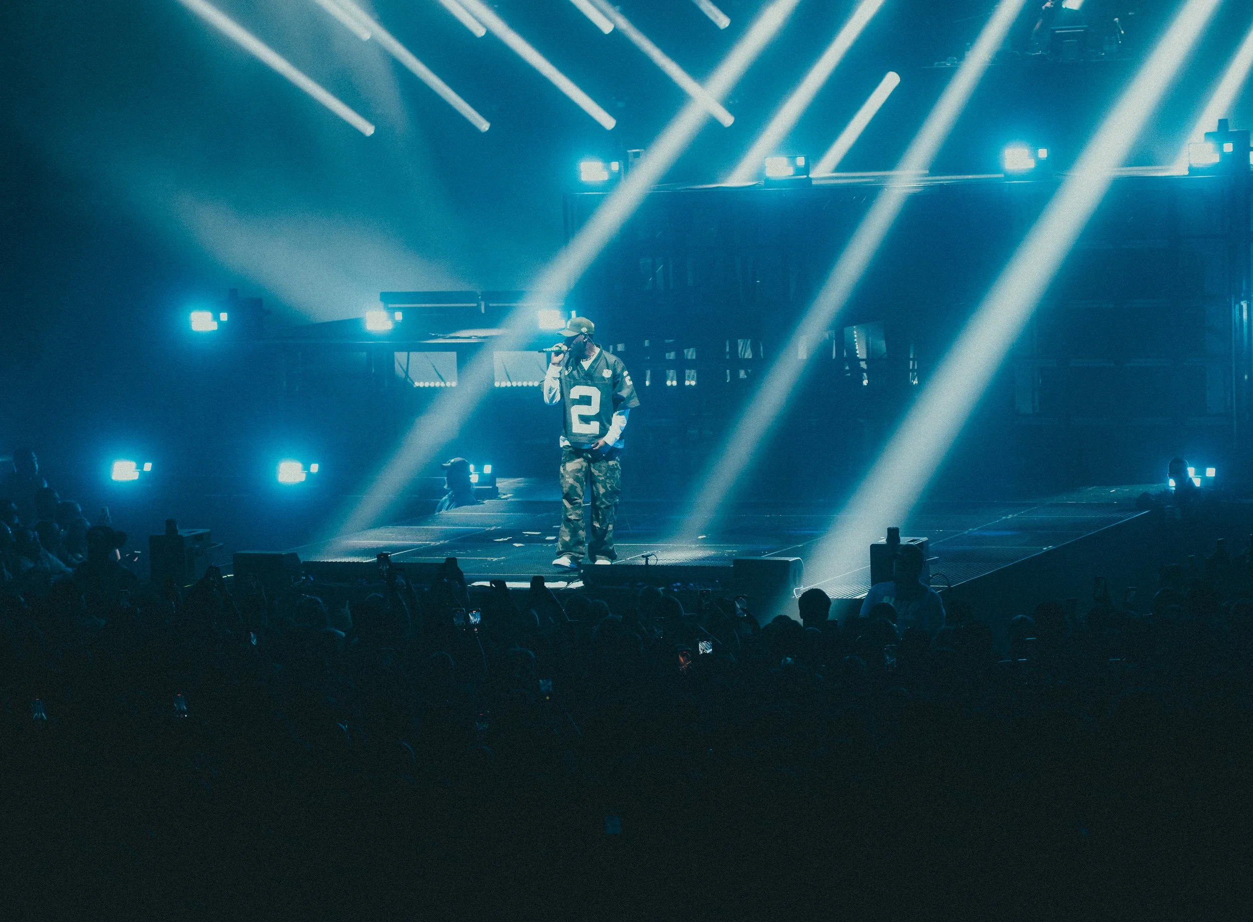 Un artiste de rap en costume camouflage sur scène, tenant un micro, avec des éclairages bleus et blancs, lumière intense, public devant.