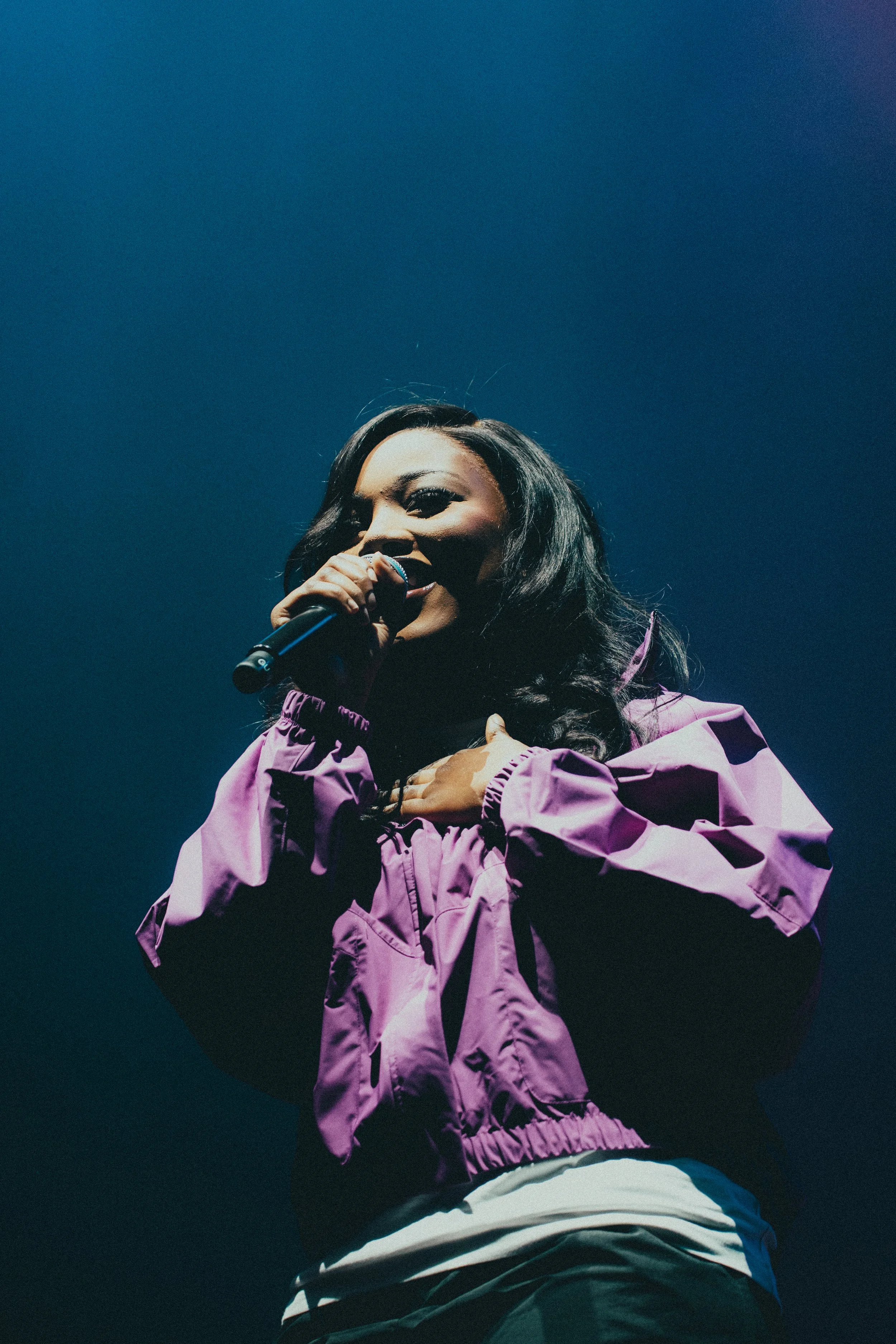 Une chanteuse noire avec de longs cheveux noirs, portant une veste violette, chante dans un micro sur scène avec un fond bleu.