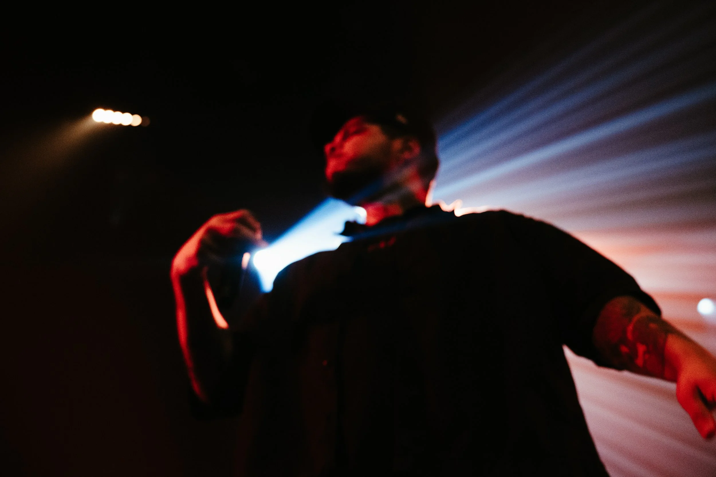 Une personne debout dans une pièce sombre avec des lumières colorées derrière elle, portant des lunettes de soleil et une chemise noire.