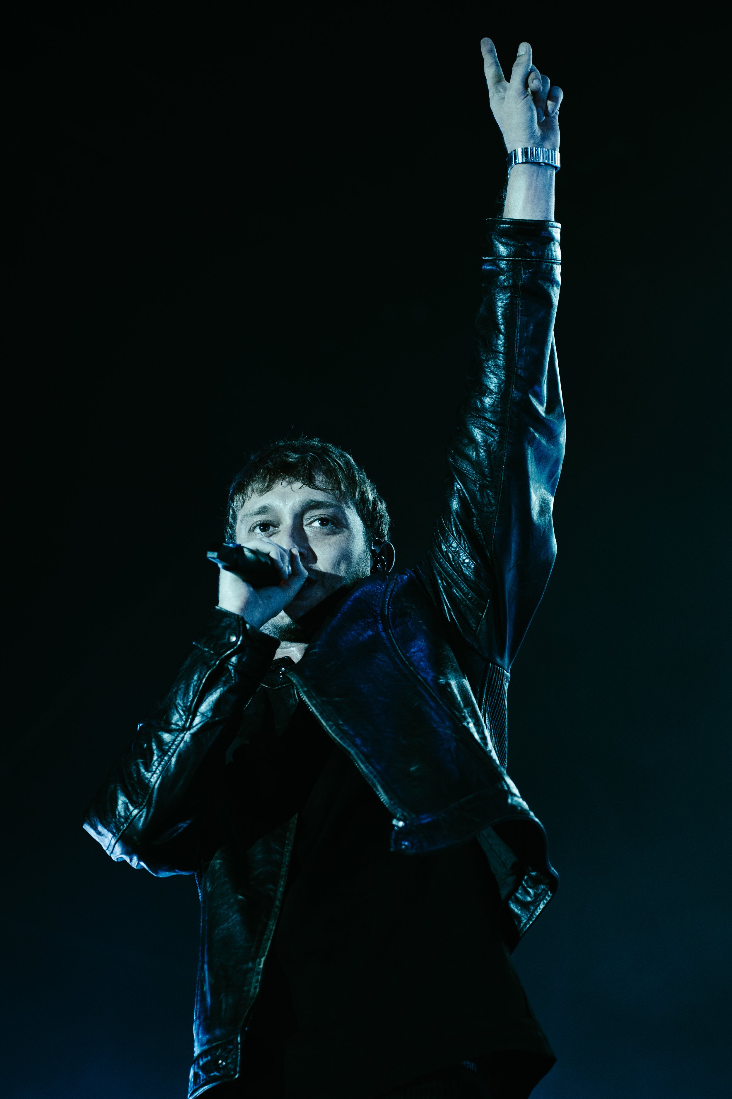Un homme chante en tenant un microphone avec un bras levé, portant une veste en cuir noire, dans un environnement sombre et éclairé par une lumière bleue.