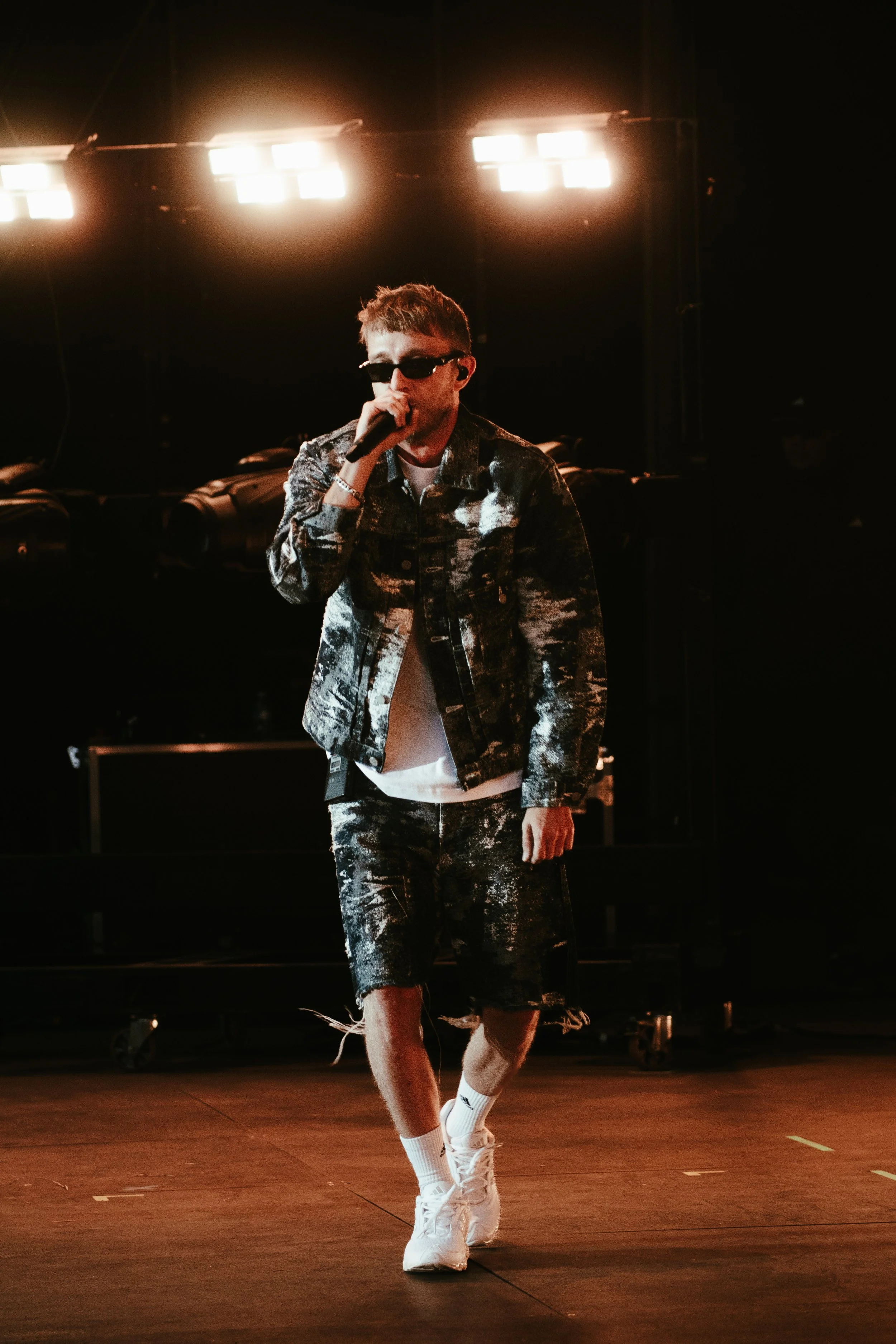 Jeune homme portant des vêtements décontractés avec un motif effet peinture, chaussures blanches, lunettes de soleil et un micro, en train de chanter ou rapper sur scène avec un fond de lumières de scène