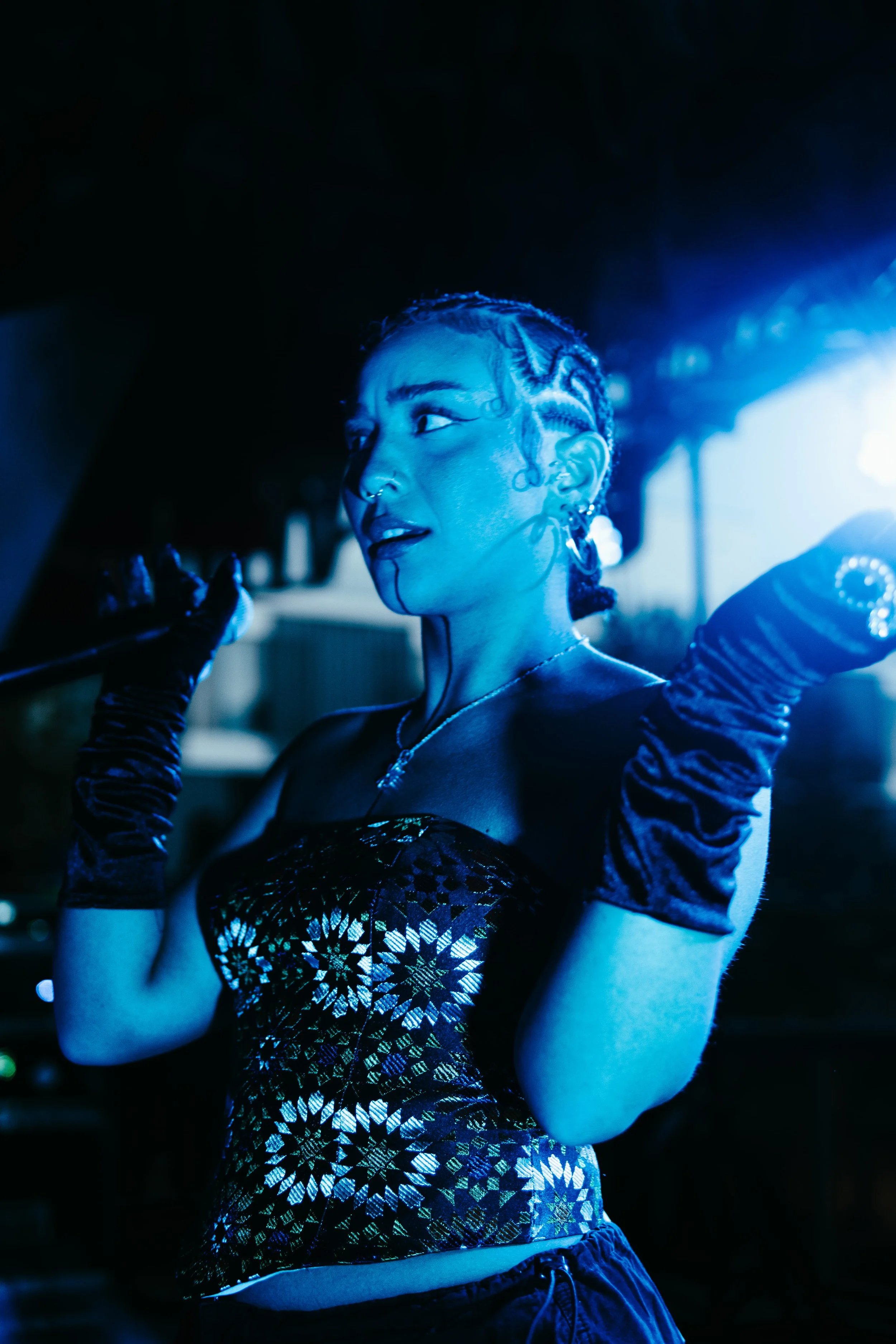Une jeune femme avec des cheveux tressés et des piercings, portant une robe à motifs dorés, des gants en velours, et un collier, dans un environnement faiblement éclairé avec une lumière bleue.