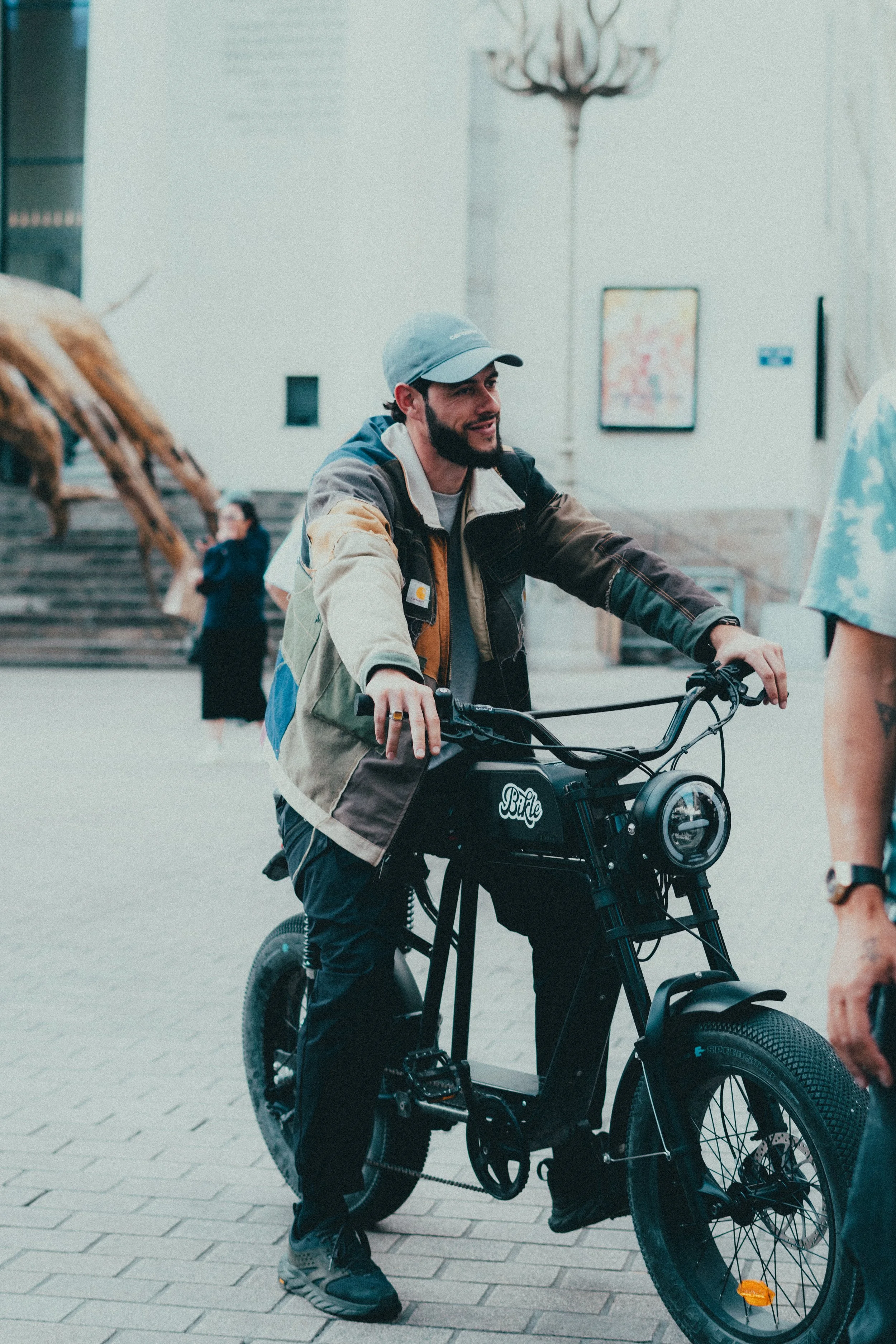 Un homme souriant en veste colorée, portant une casquette, monte dans un vélo électrique noir dans un espace intérieur avec des escaliers en arrière-plan et un éclairage de chandelier.