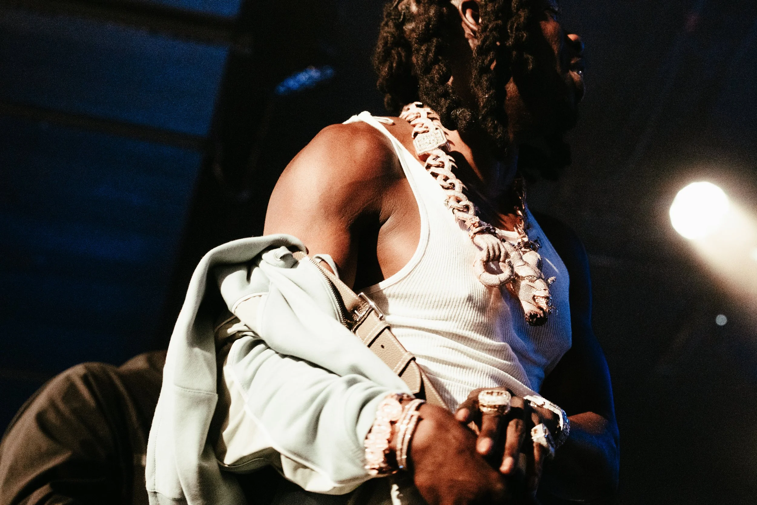 Un homme portant plusieurs bijoux, des vêtements décontractés, dans un lieu sombre avec une lumière vive derrière lui.