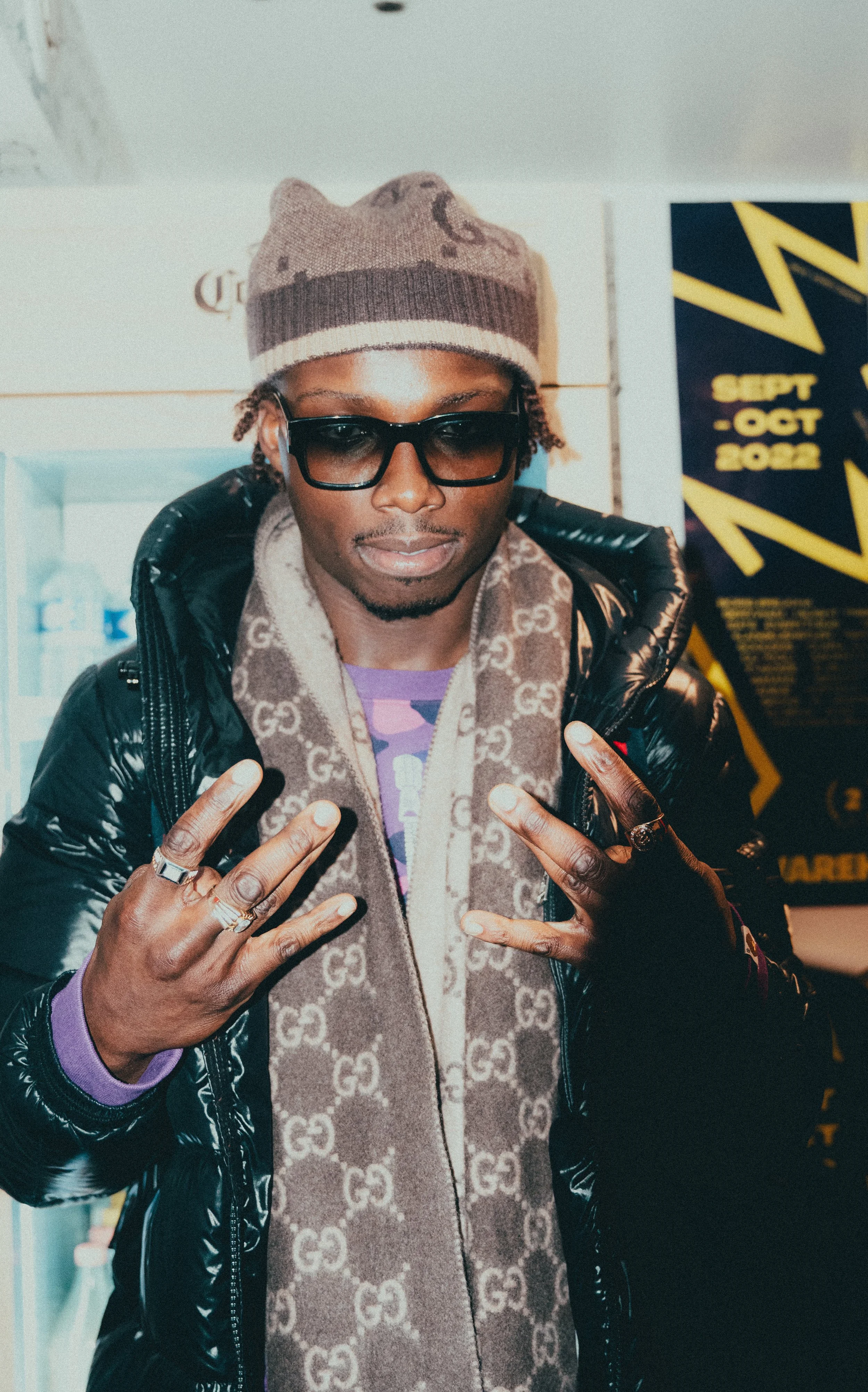 Un homme portant un bonnet marron, des lunettes de soleil noires, un manteau en cuir noir, un foulard à logo, et plusieurs bagues, faisant un geste avec ses mains dans un environnement urbain.