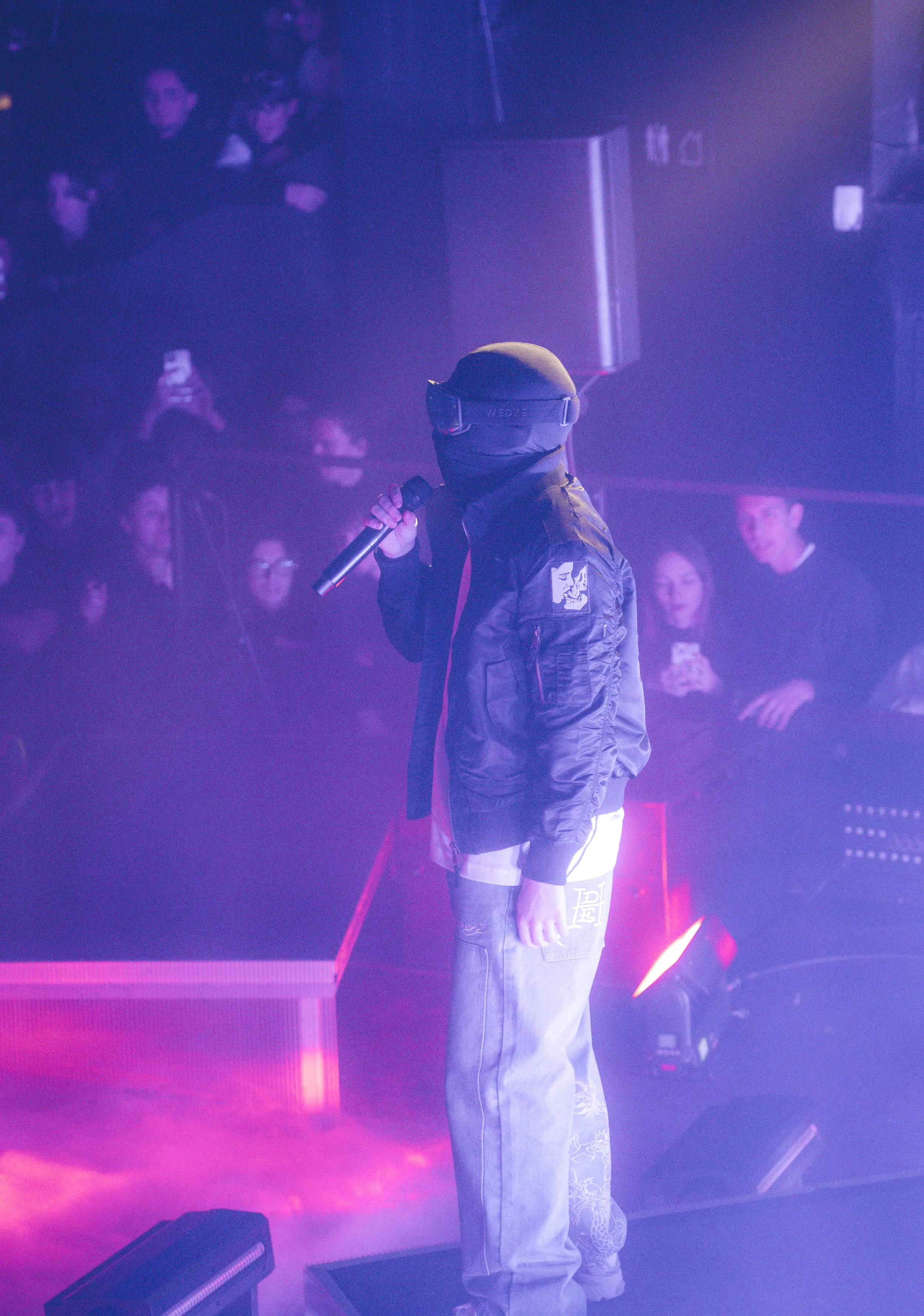 Personne portant un casque, une veste et tenant un microphone lors d'une performance sur scène avec un public en arrière-plan.