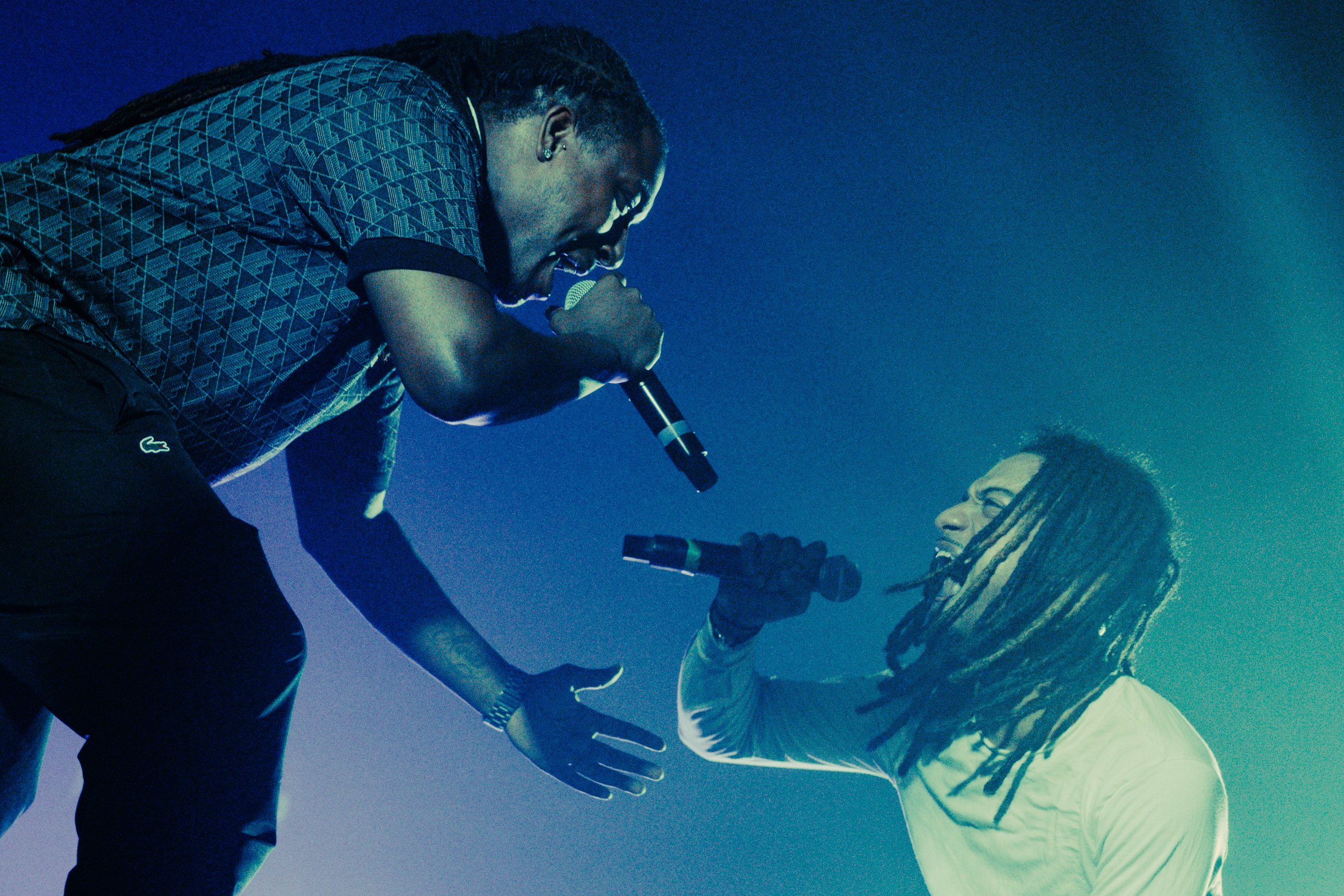 Deux hommes avec des microphones, l'un debout et l'autre à genoux, parlant ou chantant lors d'une performance ou d'un concert.