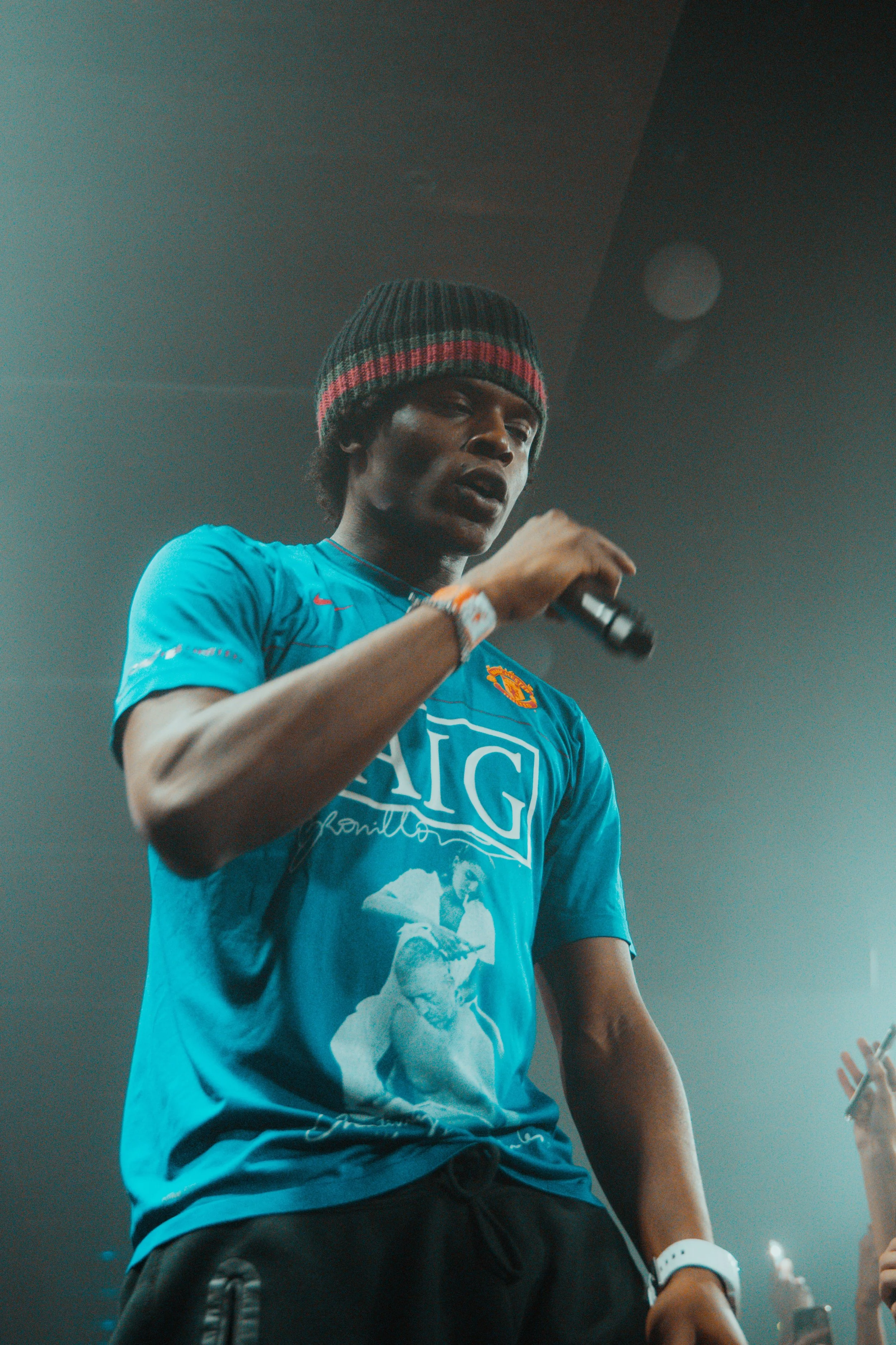 Un homme portant un bonnet noir à rayures rouges et un t-shirt bleu avec une image et du texte, tenant un microphone lors d'une prestation ou d'un discours.