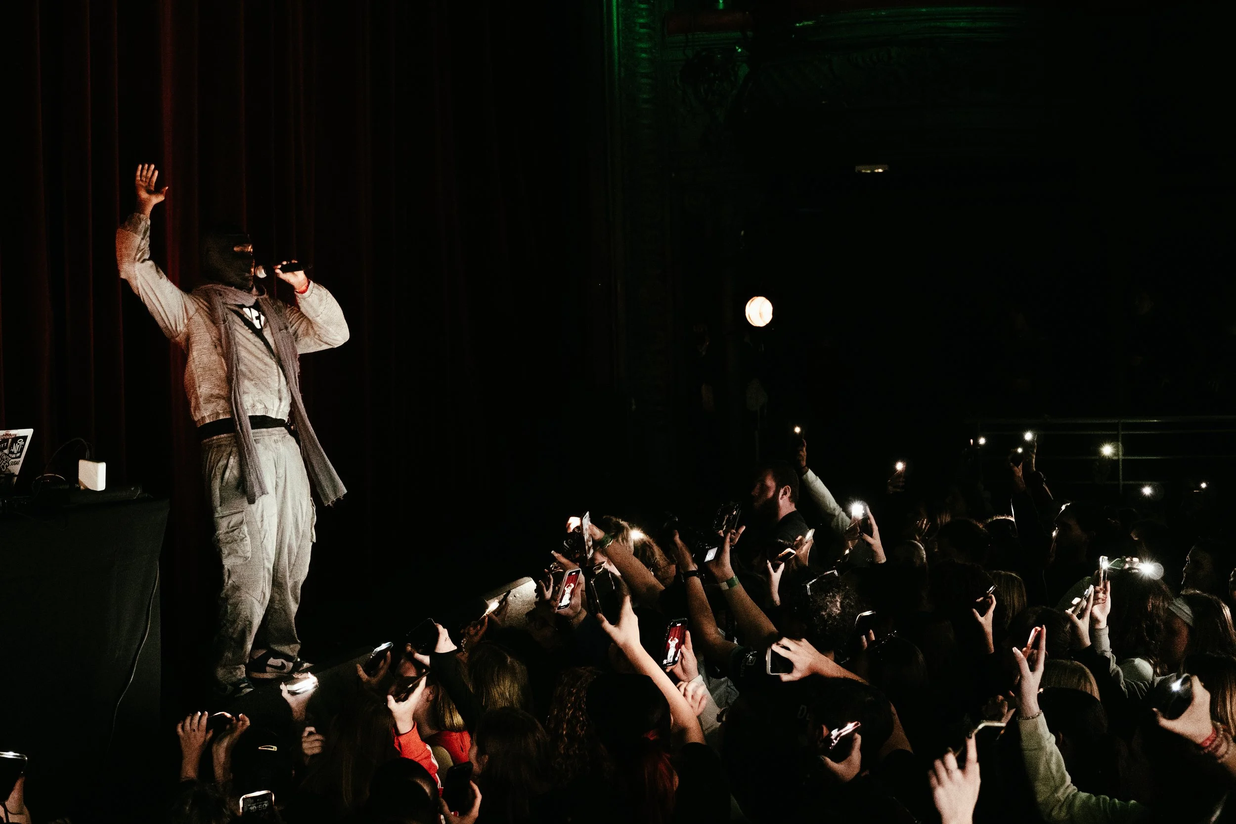 Un artiste masqué sur scène lors d'un concert, entouré d'un public qui prend des photos avec leurs téléphones portables.