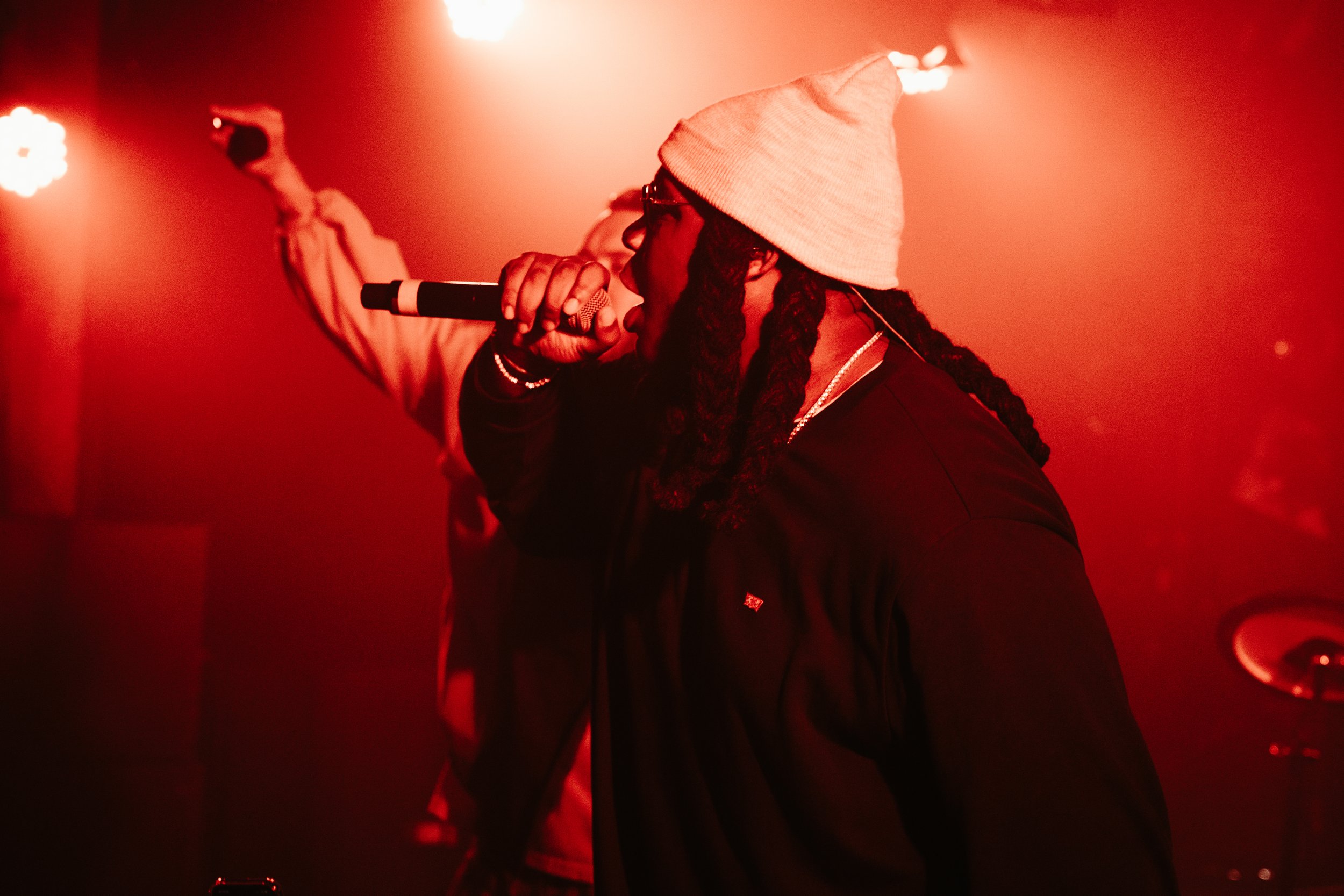 Un artiste avec des dreadlocks portant une casquette beige et des lunettes, chantant dans un microphone sous un éclairage rouge lors d'une performance.