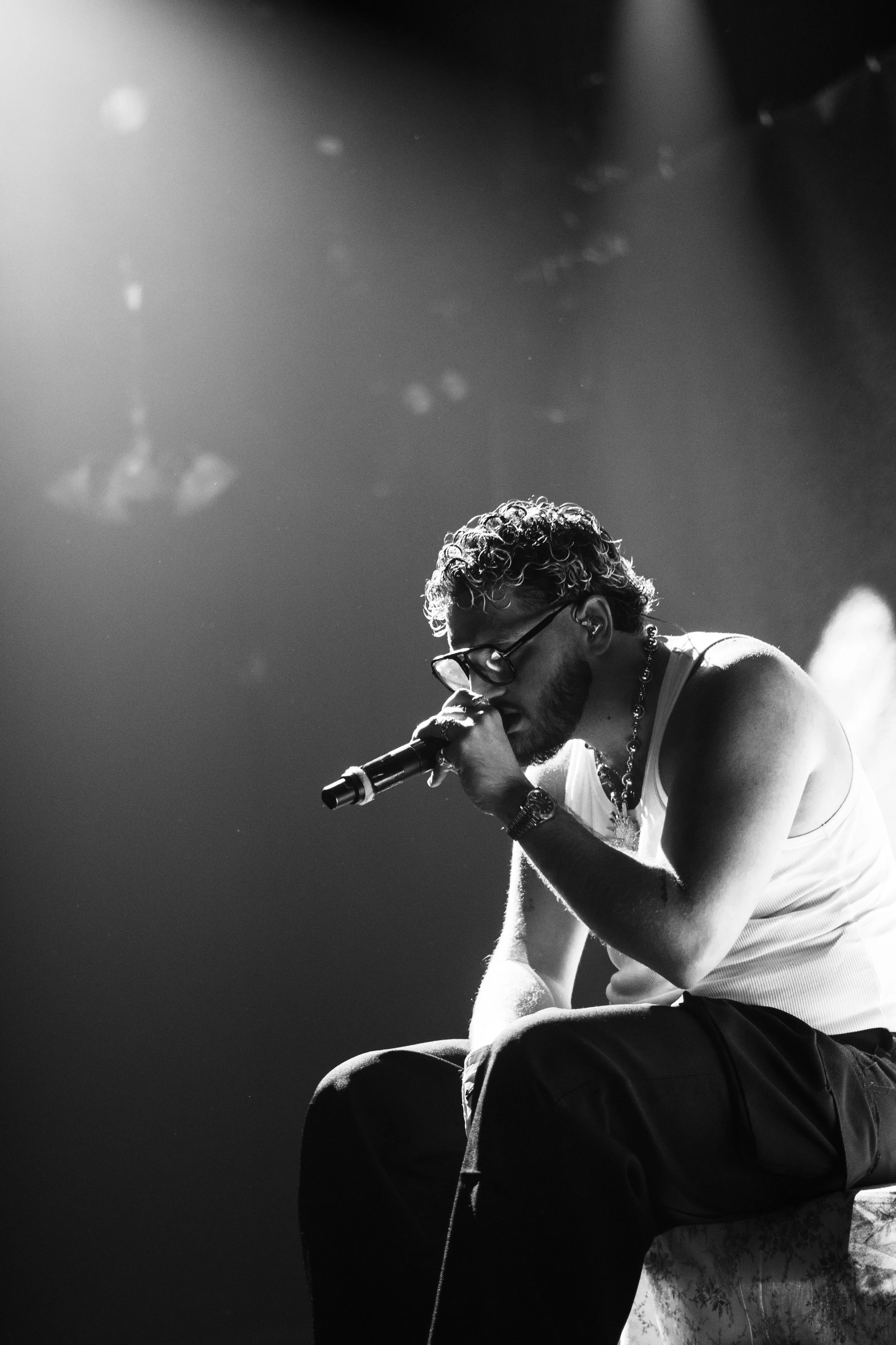 Un homme assis en train de chanter ou de parler dans un microphone sur scène, en noir et blanc, portant un t-shirt sans manches, des lunettes et un collier.