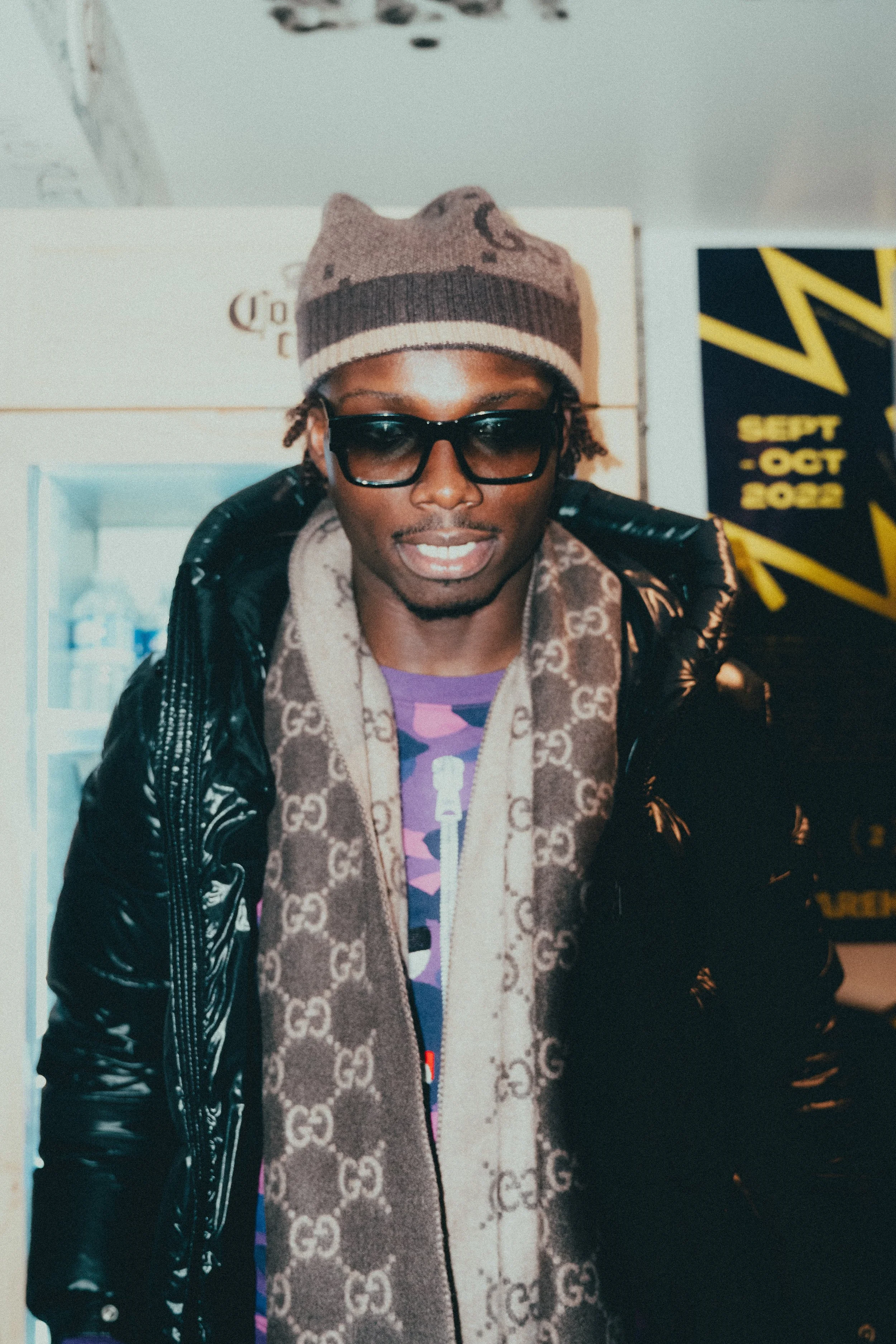 Jeune homme portant un bonnet, des lunettes de soleil noires, un manteau en tissu brillant, une écharpe avec le logo Gucci, et un t-shirt coloré, à l'intérieur, avec un panneau en arrière-plan indiquant des dates en octobre 2022.