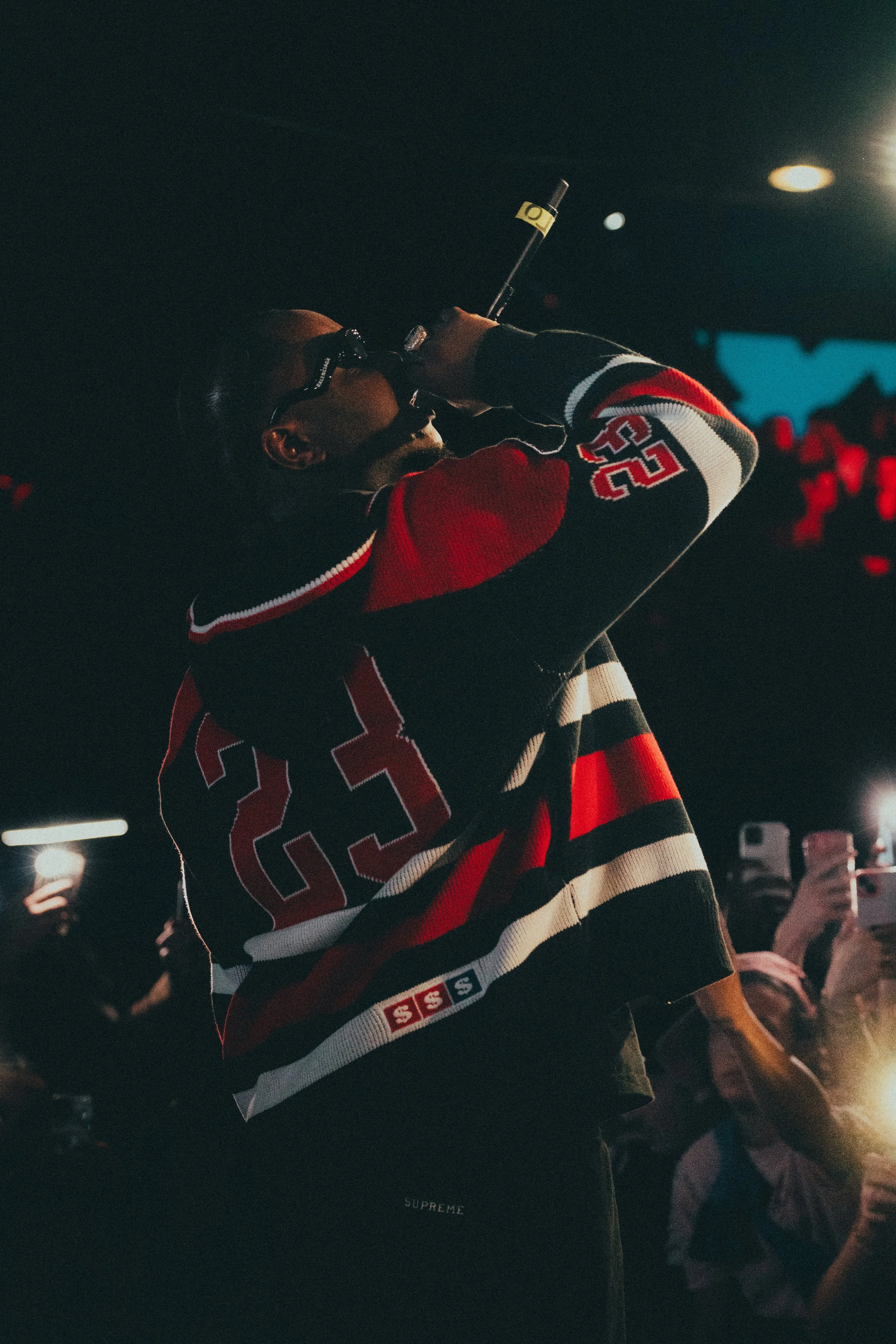 Un homme chante ou parle dans un microphone lors d'un événement ou concert, portant un maillot de hockey noir, rouge, blanc avec le numéro 25. Il porte des lunettes et se tient dans une salle sombre avec une foule en arrière-plan.