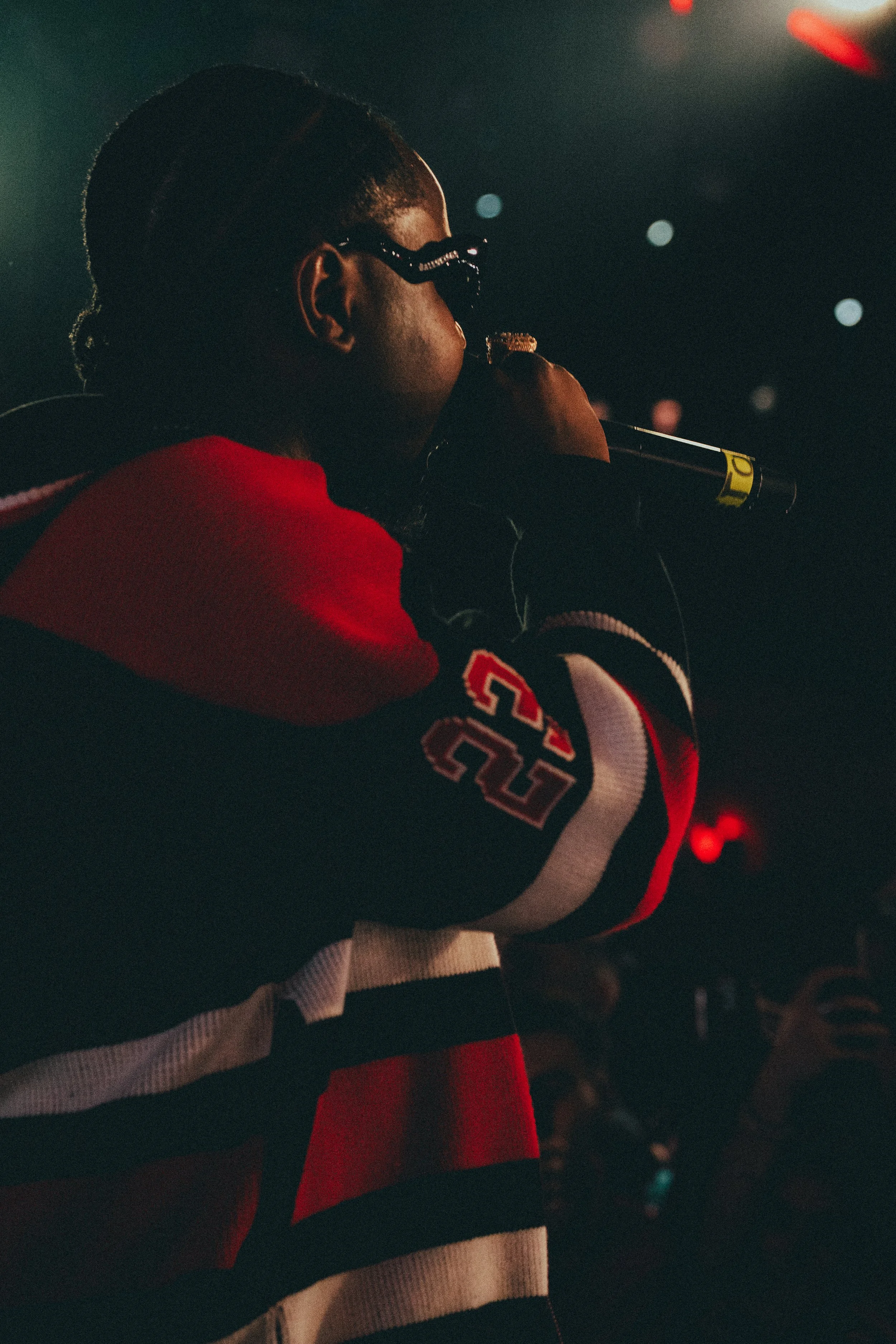 Une personne portant des lunettes de soleil, vêtue d'un pull à rayures rouge, noir et blanc portant le numéro 23, tenant un microphone dans un environnement sombre avec des lumières diffuses.