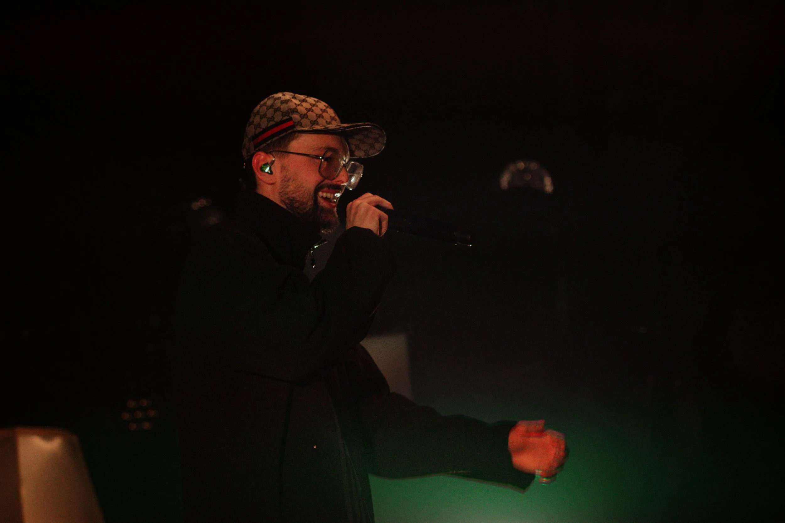 Un homme chante ou parle dans un micro lors d'une performance ou d'un événement dans un environnement sombre.