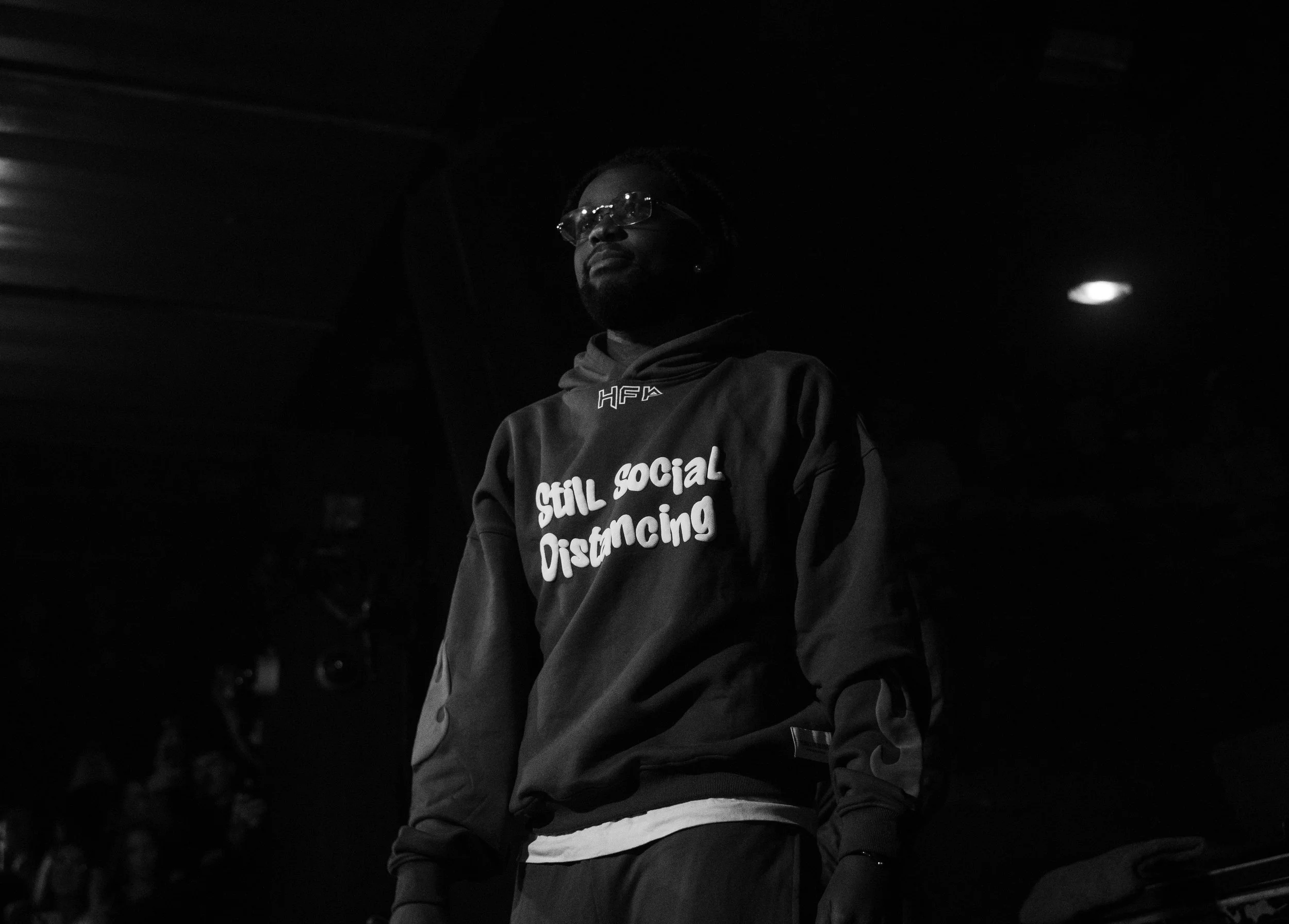 Un homme porte un sweat à capuche avec le texte 'Still Social Distancing' en blanc, dans une ambiance sombre et en noir et blanc.