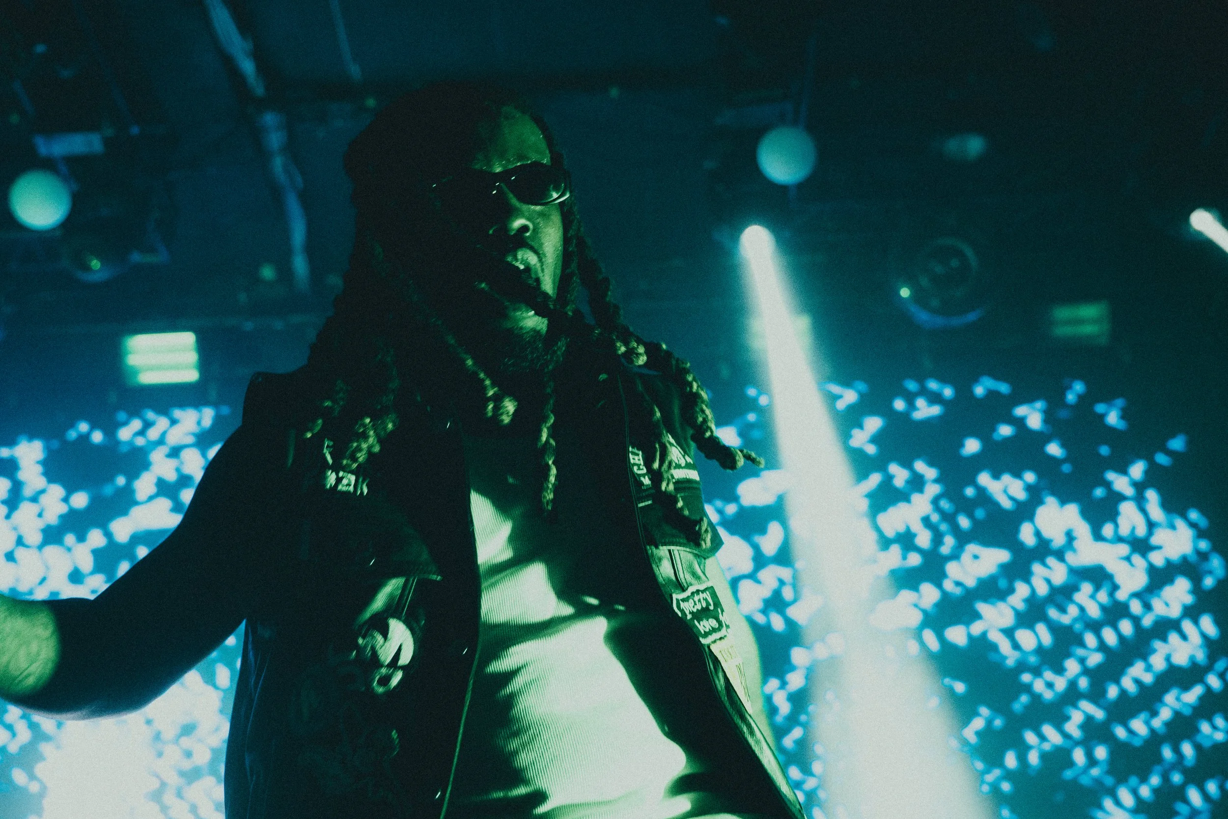 Un artiste avec des dreadlocks, portant des lunettes de soleil et un blouson à patches lumineux, performe sur scène avec des lumières colorées et un écran vidéo en arrière-plan.