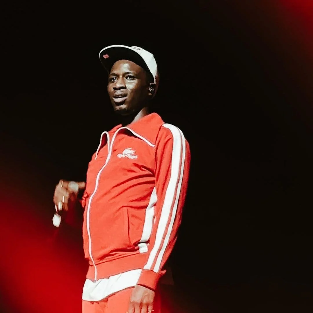 ORMAZ avec un casque, portant une veste de sport rouge avec des bandes blanches, souriant, en arrière-plan sombre.