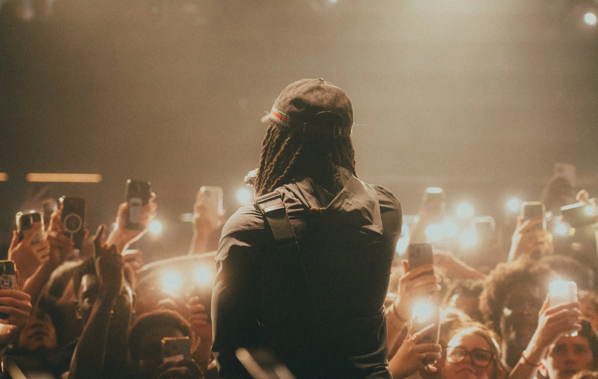 Un artiste ou un intervenant avec des dreadlocks, portant une casquette et un vestiaire sombre, se tient devant une foule lors d'un concert ou d'un événement. La scène est éclairée par de nombreuses lumières et la foule tient des téléphones portables
