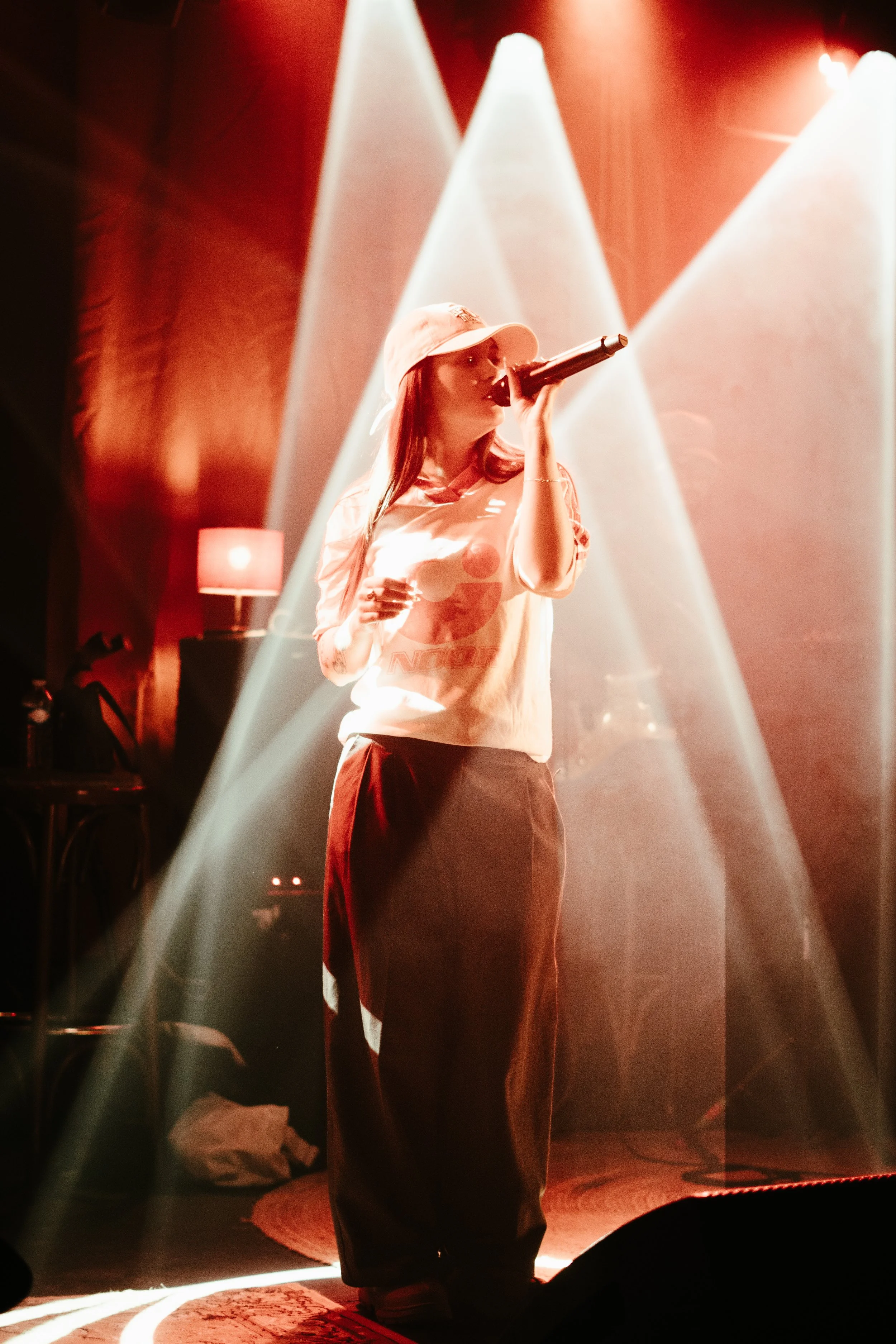 Une femme chante avec un micro sur scène éclairée par des faisceaux lumineux, ambiance de concert. Elle porte un chapeau blanc et un t-shirt blanc. L'arrière-plan est sombre avec une lampe et des sources lumineuses.
