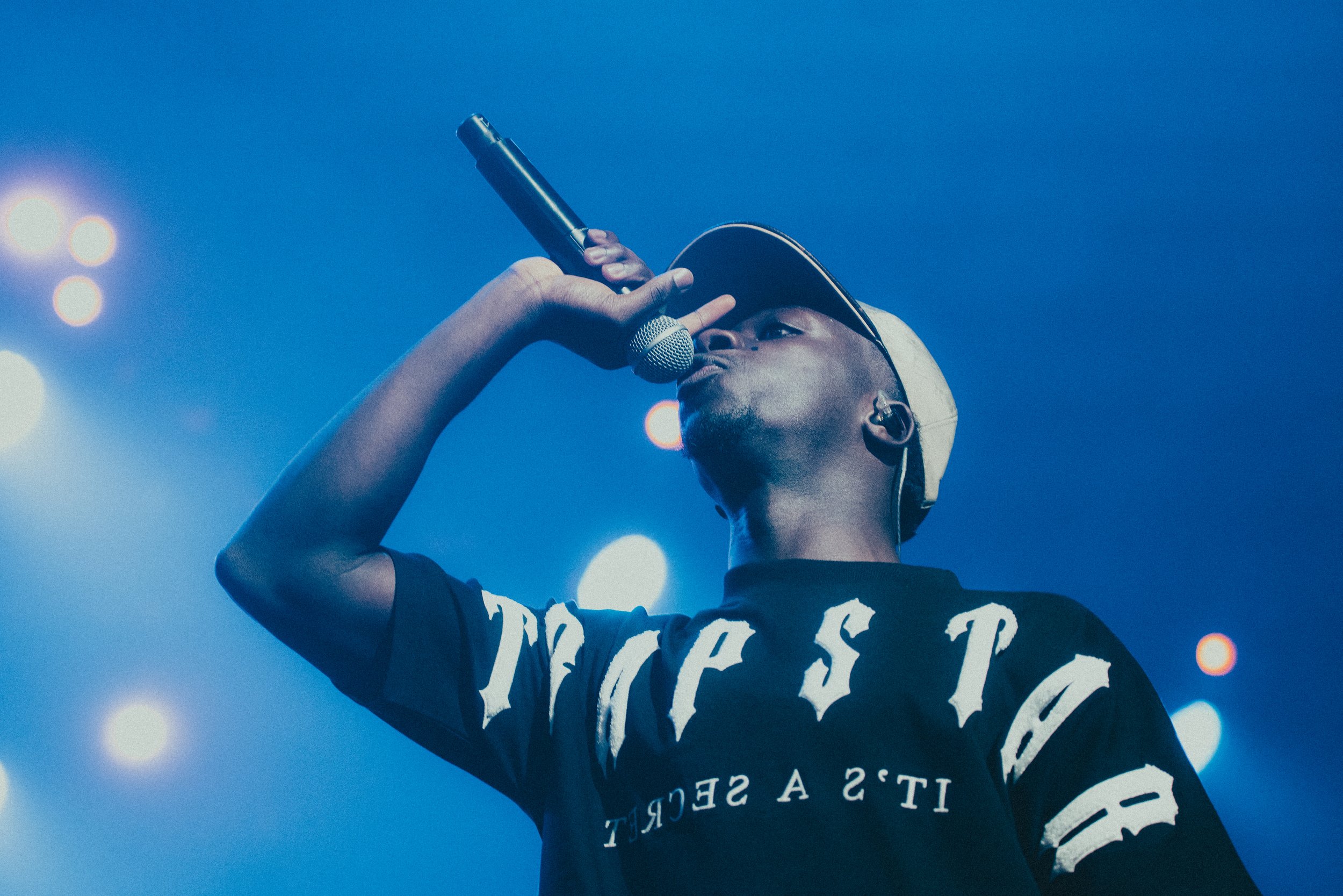 Un jeune rappeur en train de chanter ou de rapper sur scène, portant une casquette et un t-shirt noir, avec un microphone. Les lumières de scène en arrière-plan.