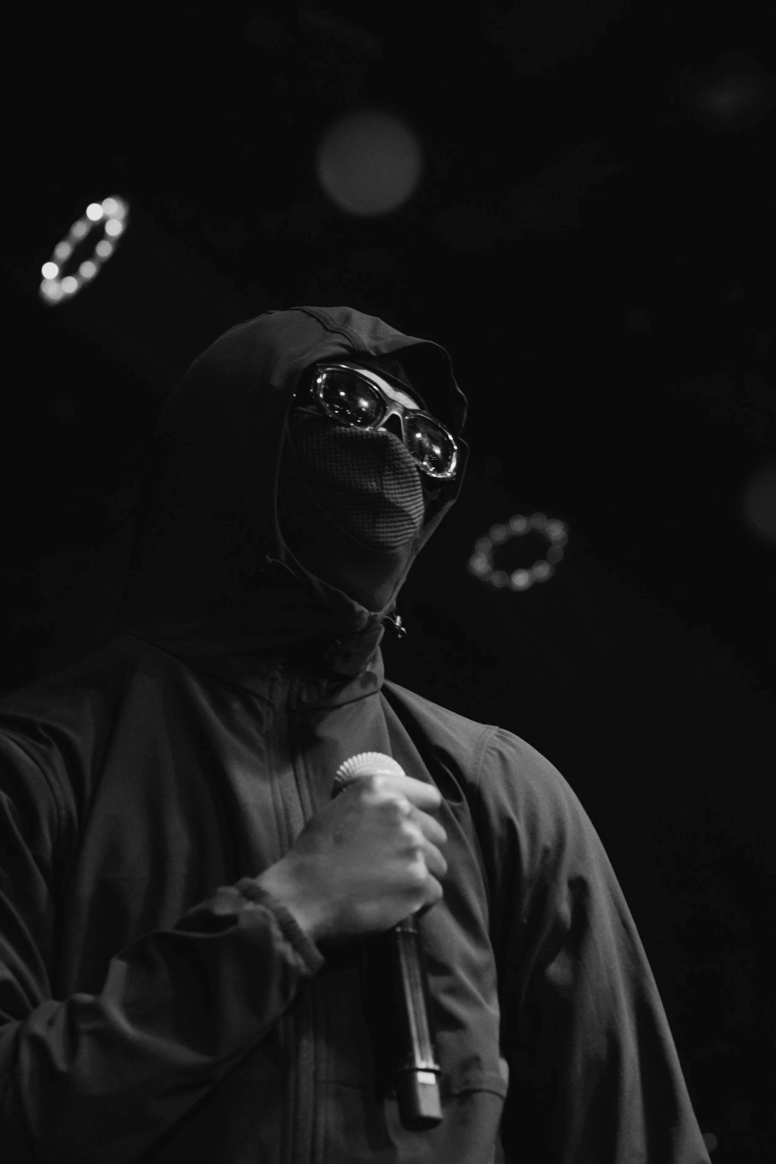 Personne portant un sweat à capuche, une paire de lunettes de protection et un masque, tenant un microphone dans une pièce sombre.