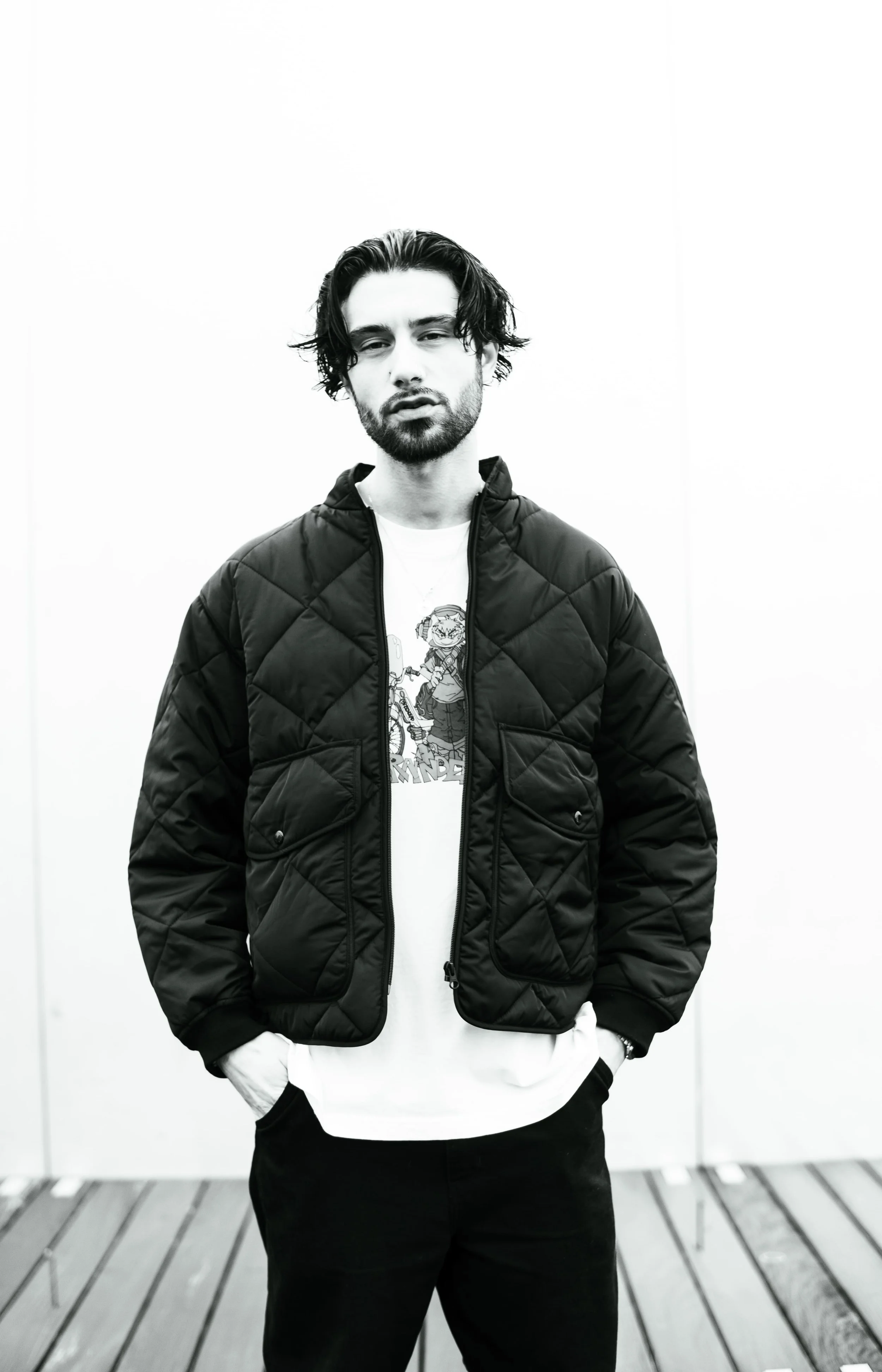 Jeune homme à l'air pensif portant une veste matelassée noire, un t-shirt blanc avec un dessin, et un pantalon noir, se tenant devant un mur blanc.