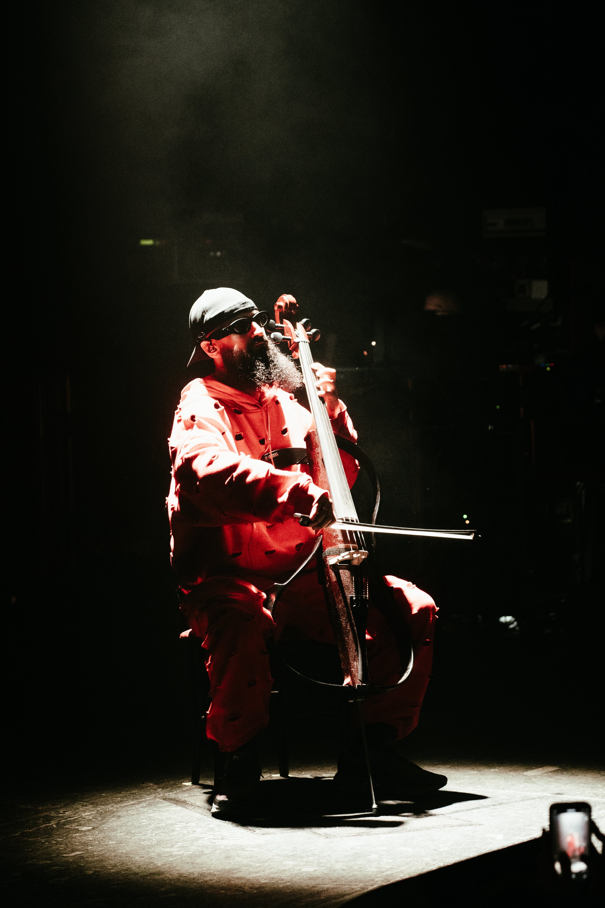 Un homme jouant du violoncelle sur scène, éclairé par une lumière tamisée, portant une casquette, des lunettes noires, une veste rouge à pois et un pantalon assorti.