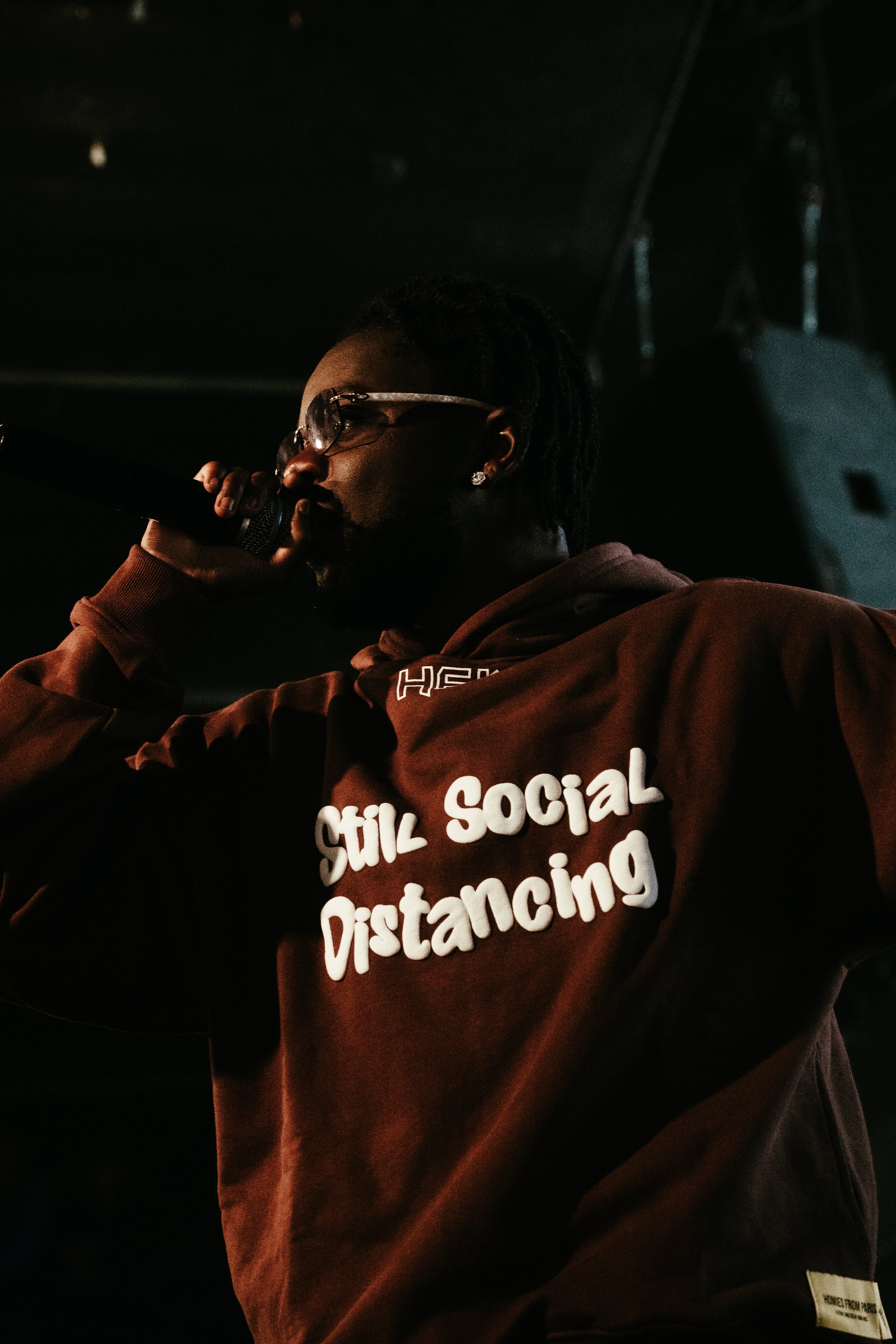 Un homme portant une veste marron avec la phrase "Still Social Distancing" écrit dessus, tenant un micro et portant des lunettes de soleil, dans un environnement sombre.