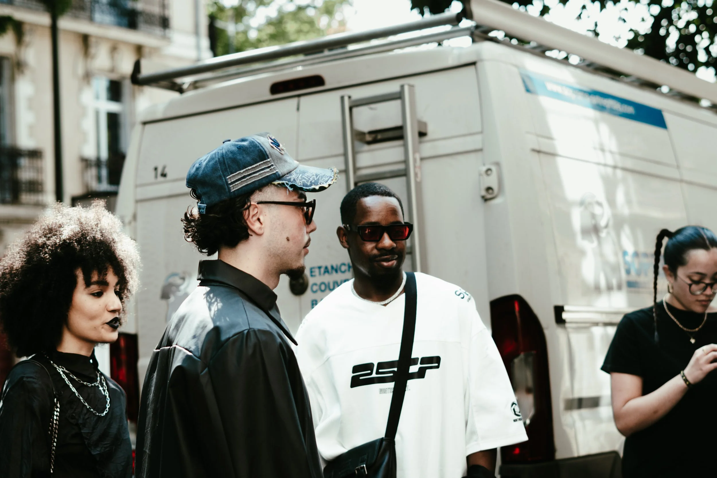 Groupe de jeunes adultes debout devant un camion blanc, portant des vêtements modernes et des accessoires, dans une rue urbaine.