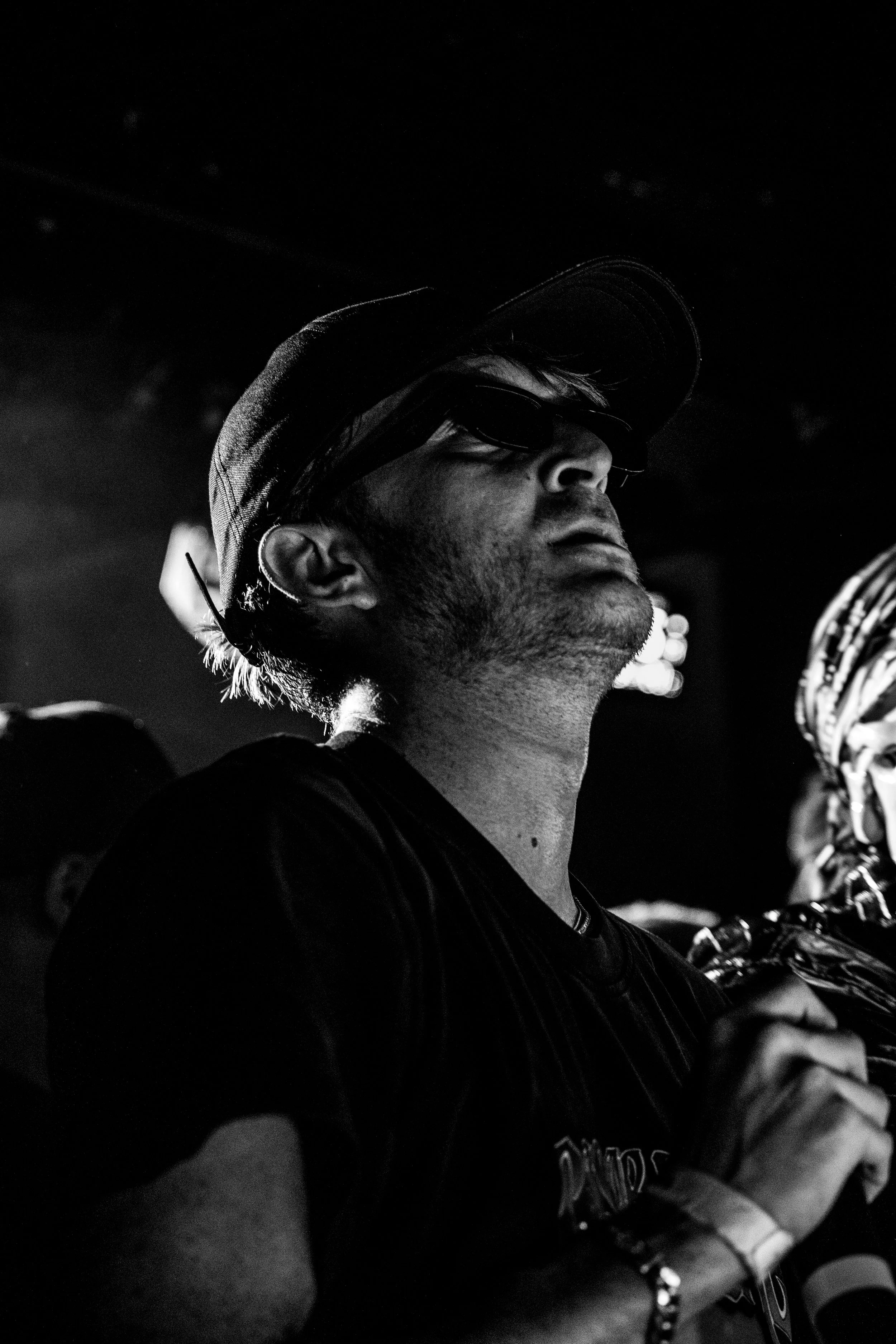 Un homme portant une casquette, des lunettes de soleil et un t-shirt noir, regardant vers le haut, en noir et blanc.