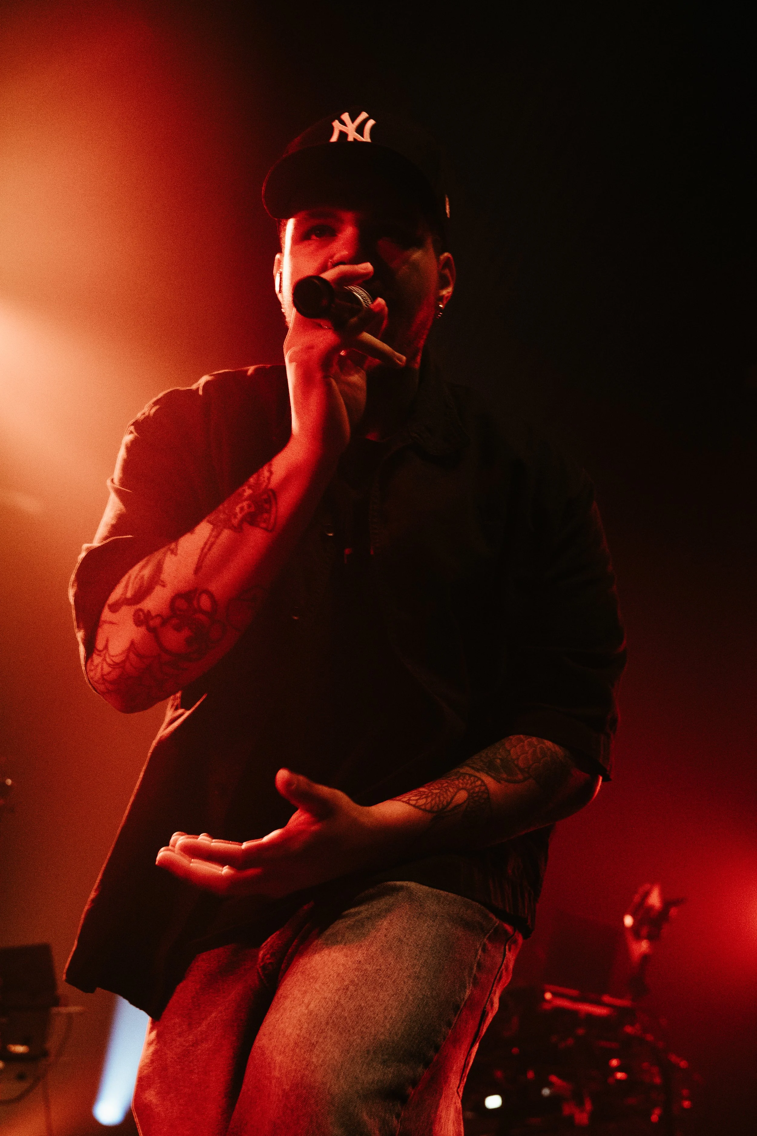 Un homme avec un tatouage au bras tenant un microphone, portant une casquette noire avec le logo NY, en train de chanter ou parler lors d'une performance sous un éclairage rouge.