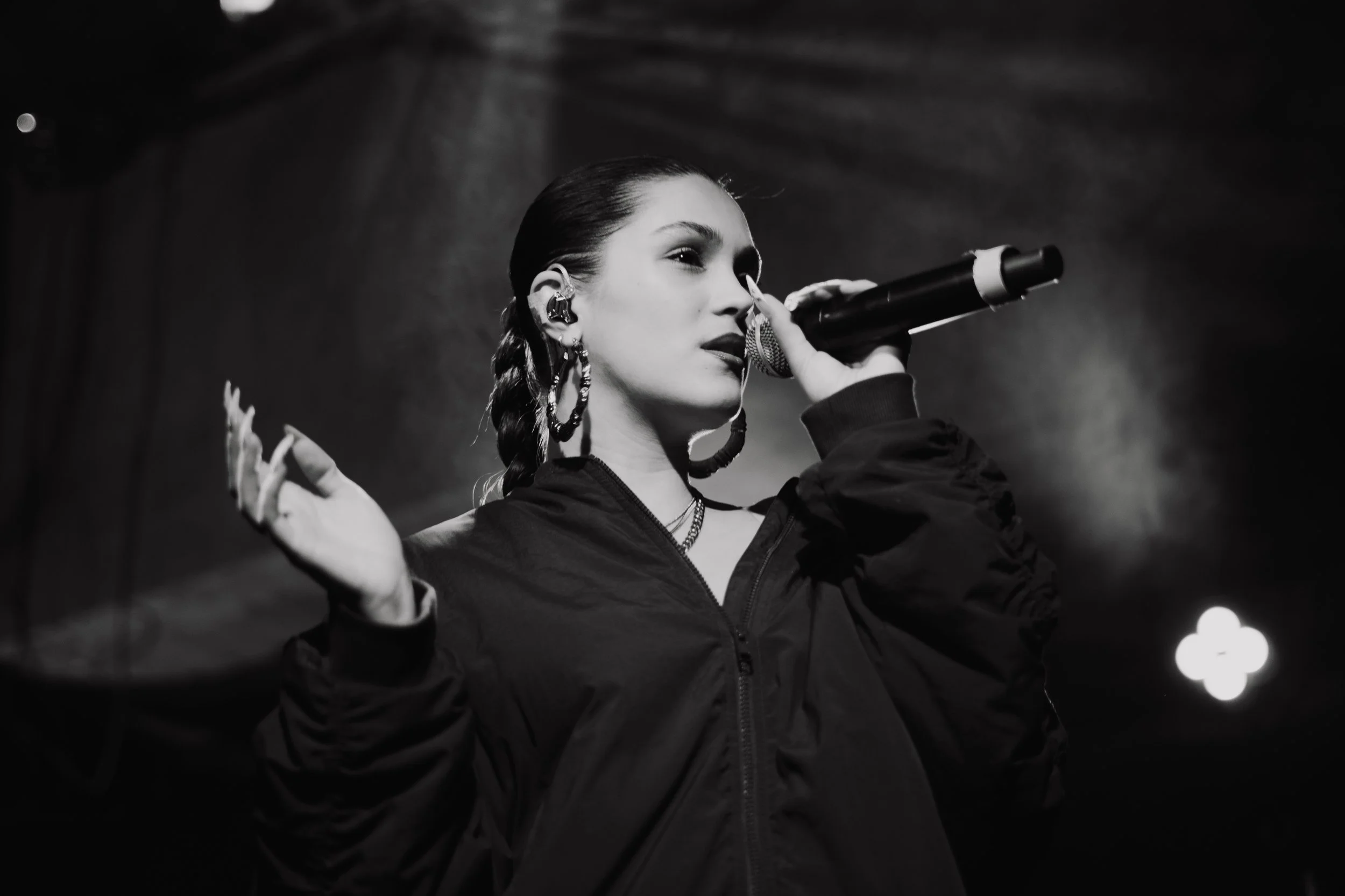 Une femme chante en noir et blanc, tenant un micro près de la bouche, avec des boucles d'oreilles et des accessoires, sur scène avec un éclairage en fond.