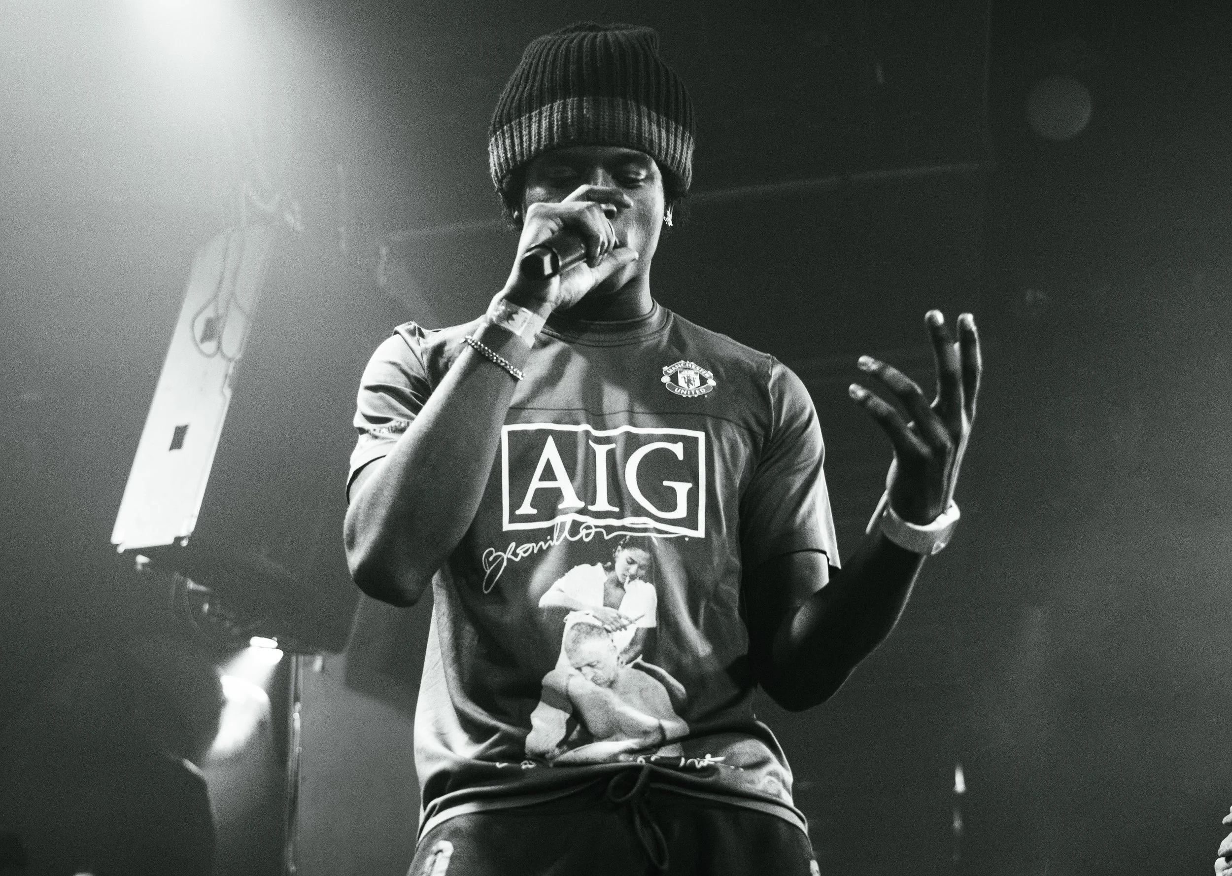 Une personne portant un t-shirt avec le logo AIG et une image d'une femme, tenant un micro, sur scène, avec un éclairage en arrière-plan. En noir et blanc.