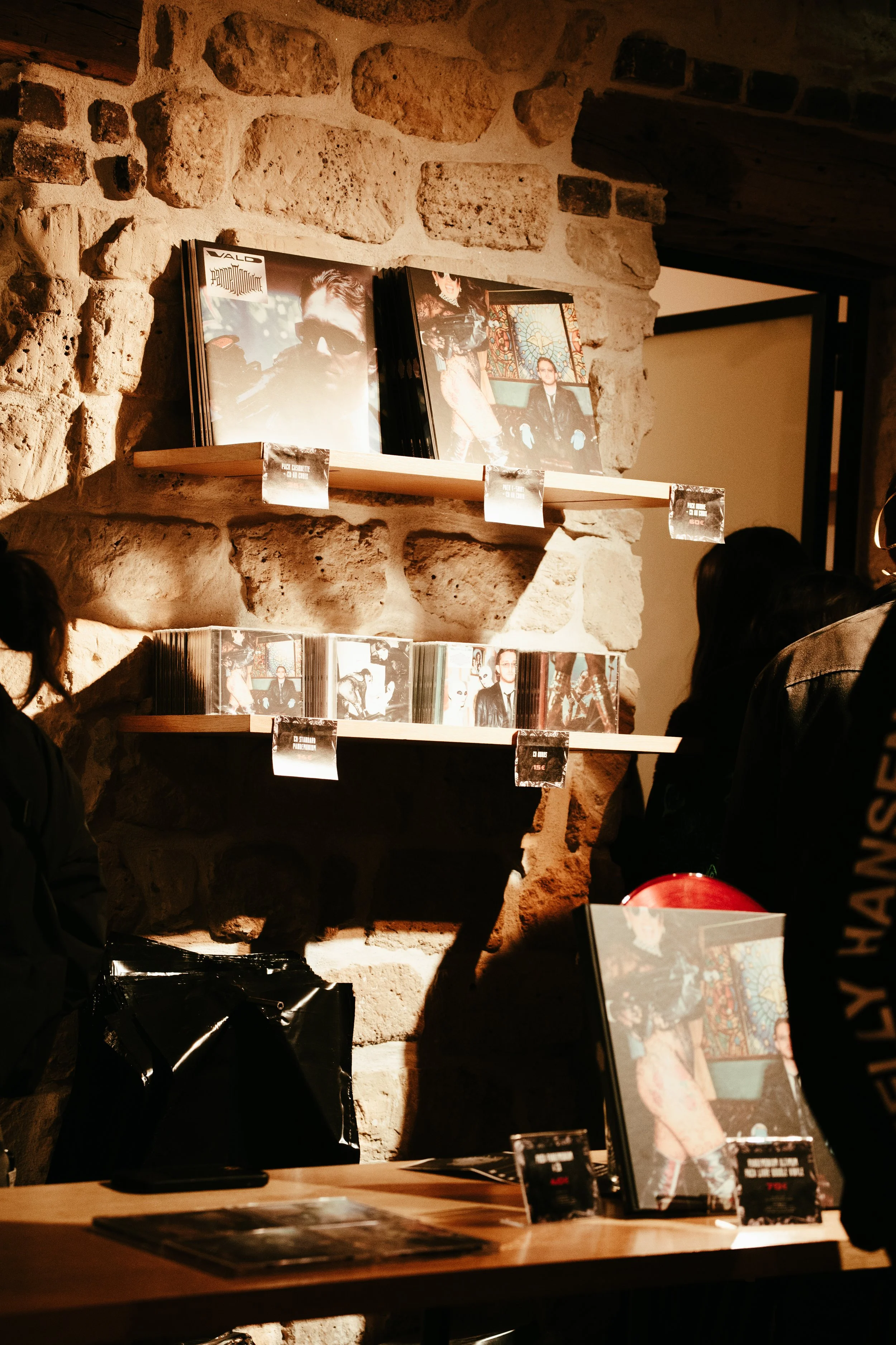 Exposition de pochettes de disques sur des étagères en bois fixées à un mur en pierre, dans un lieu probablement une boutique ou une salle d'exposition musicale, avec des personnes observant les œuvres.