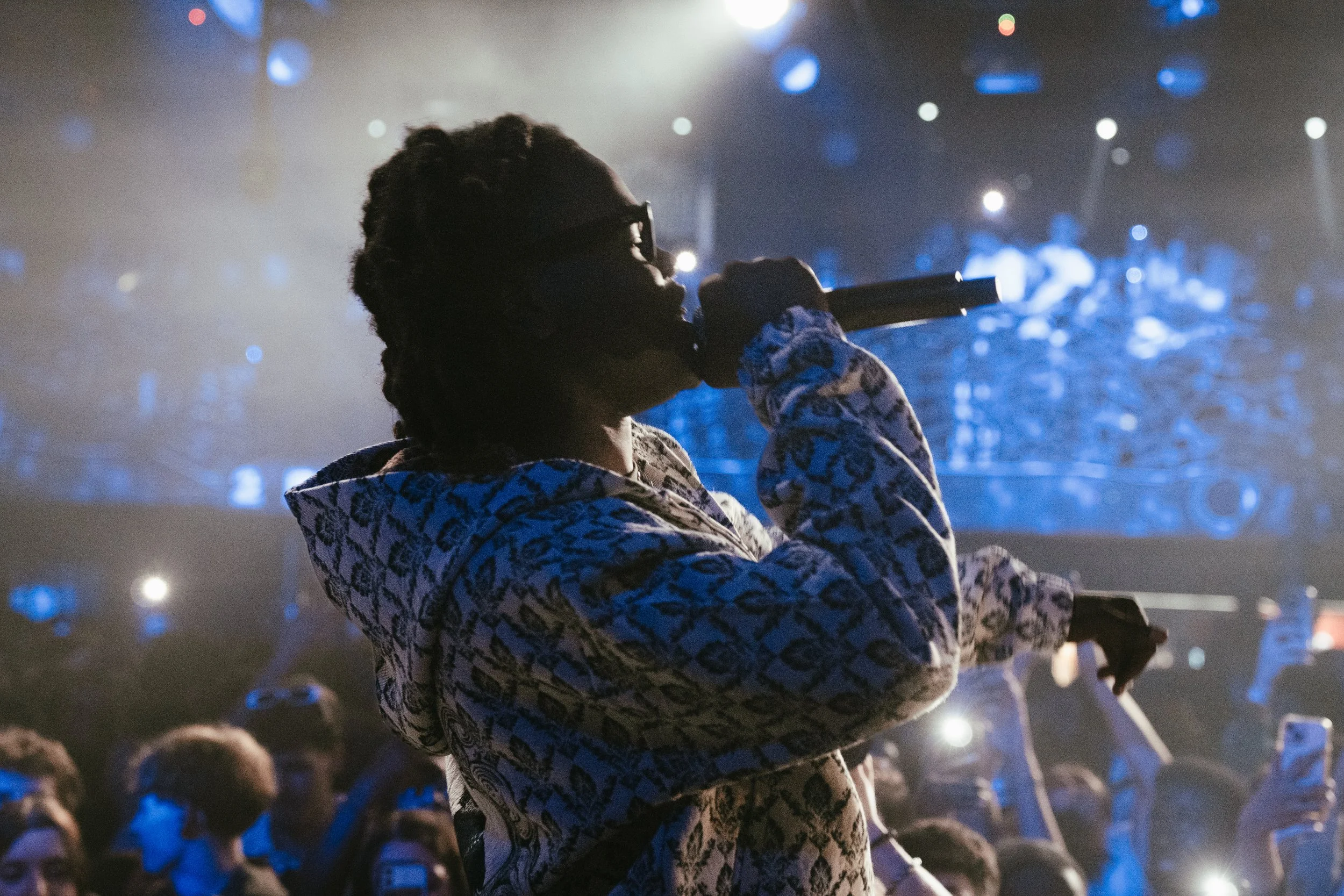 Une personne chante ou parle dans un microphone lors d'un concert, avec une foule et des lumières colorées à l'arrière-plan.