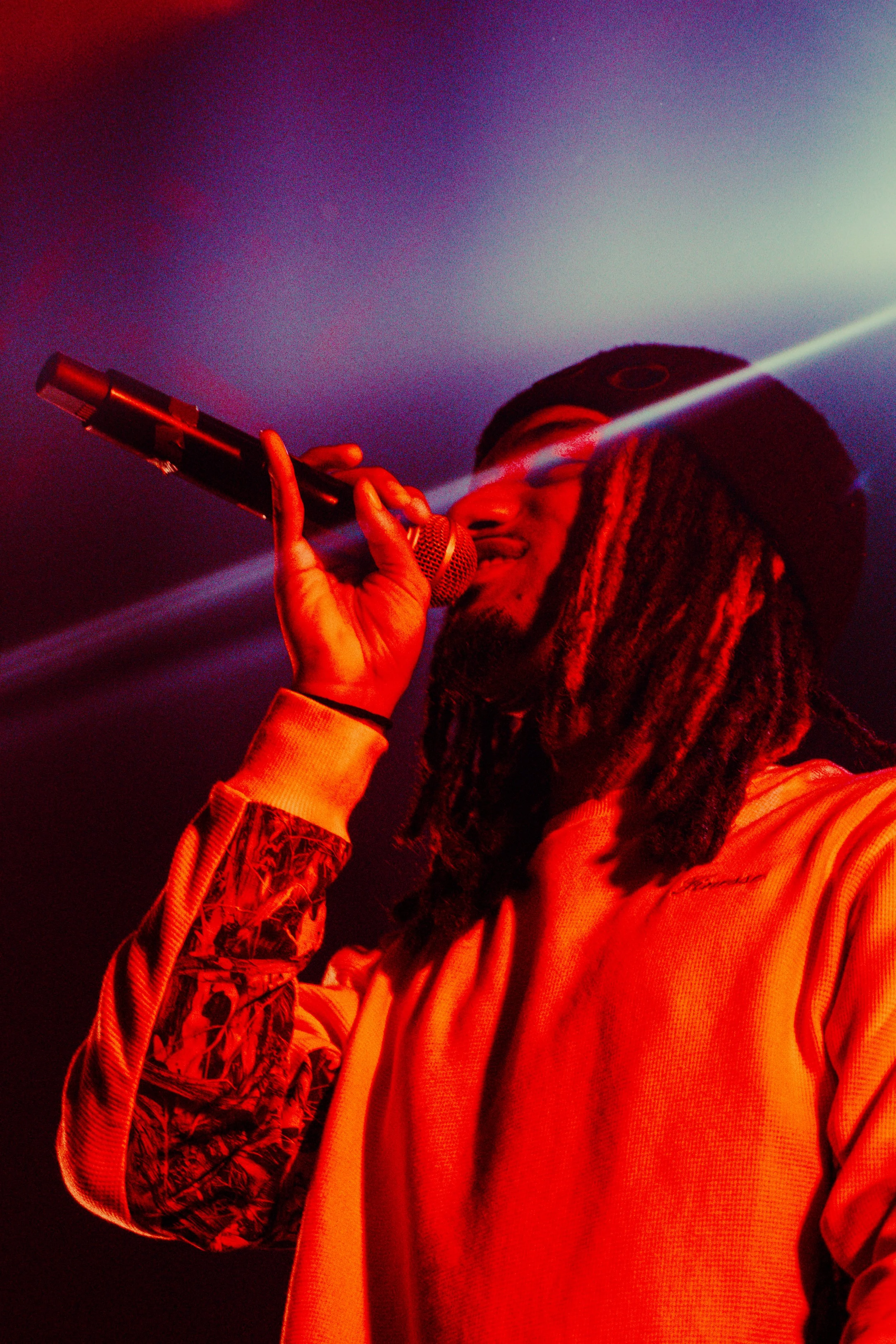 Un homme avec des dreadlocks portant un bonnet noir, chantant dans un microphone lors d'une performance. Éclairage rouge et violet dans l'ambiance.