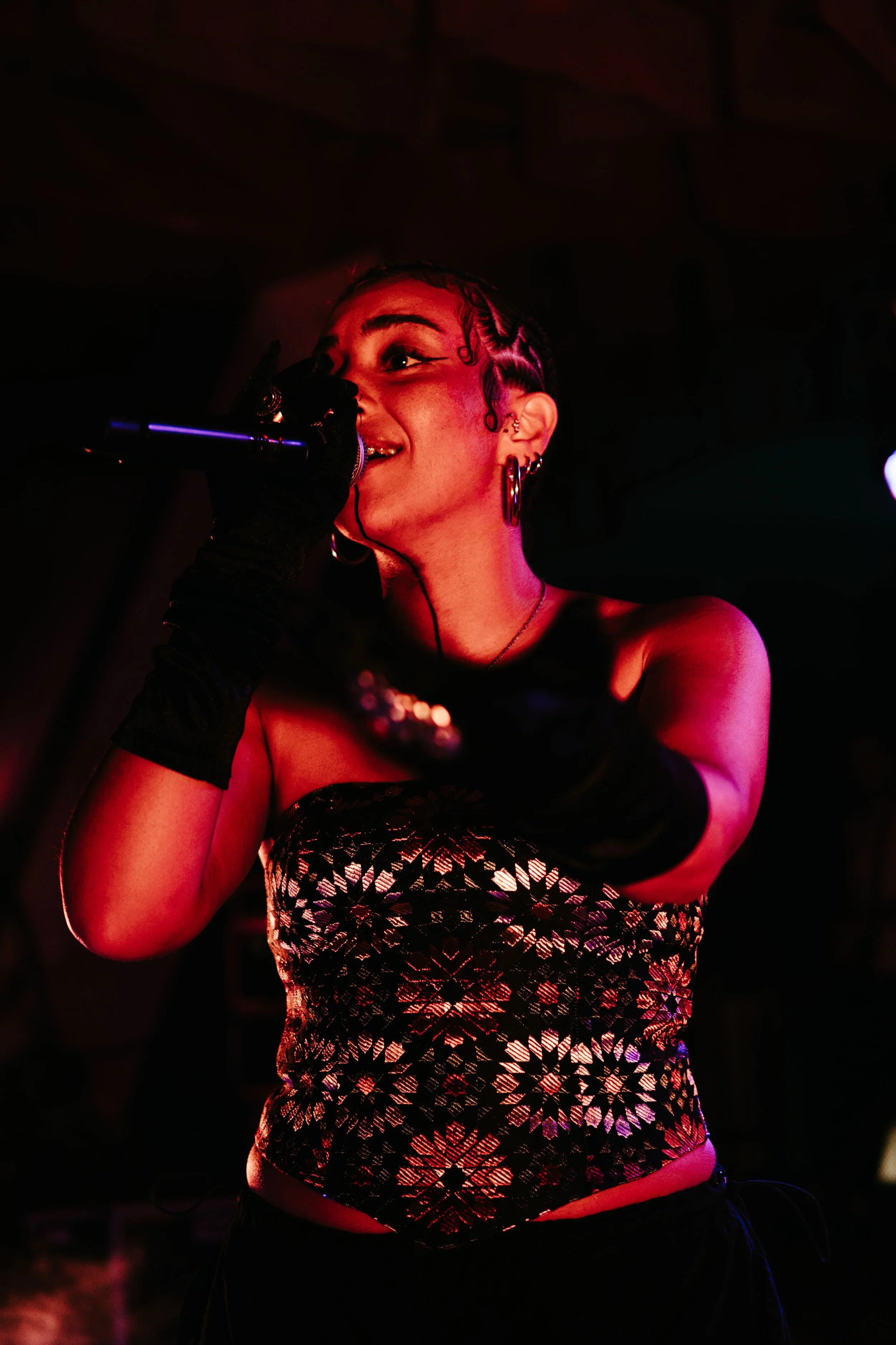 Une femme chante dans un micro avec un éclairage coloré, portant un haut noir à motifs géométriques et des gants noirs longs.
