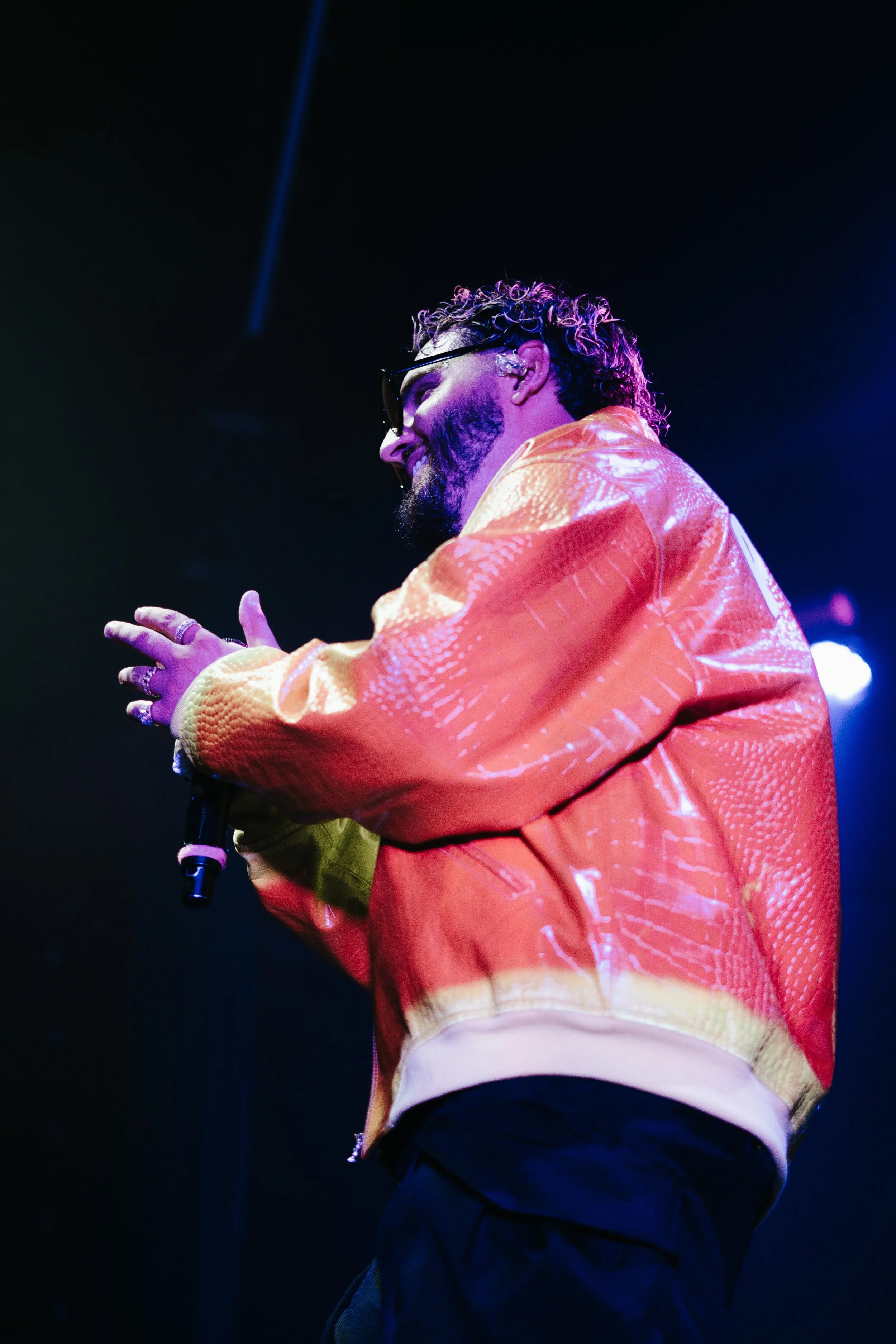Un homme avec des cheveux bouclés, portant des lunettes de soleil et une veste en cuir orange, est en train de chanter ou de parler au microphone lors d'une performance sur scène.