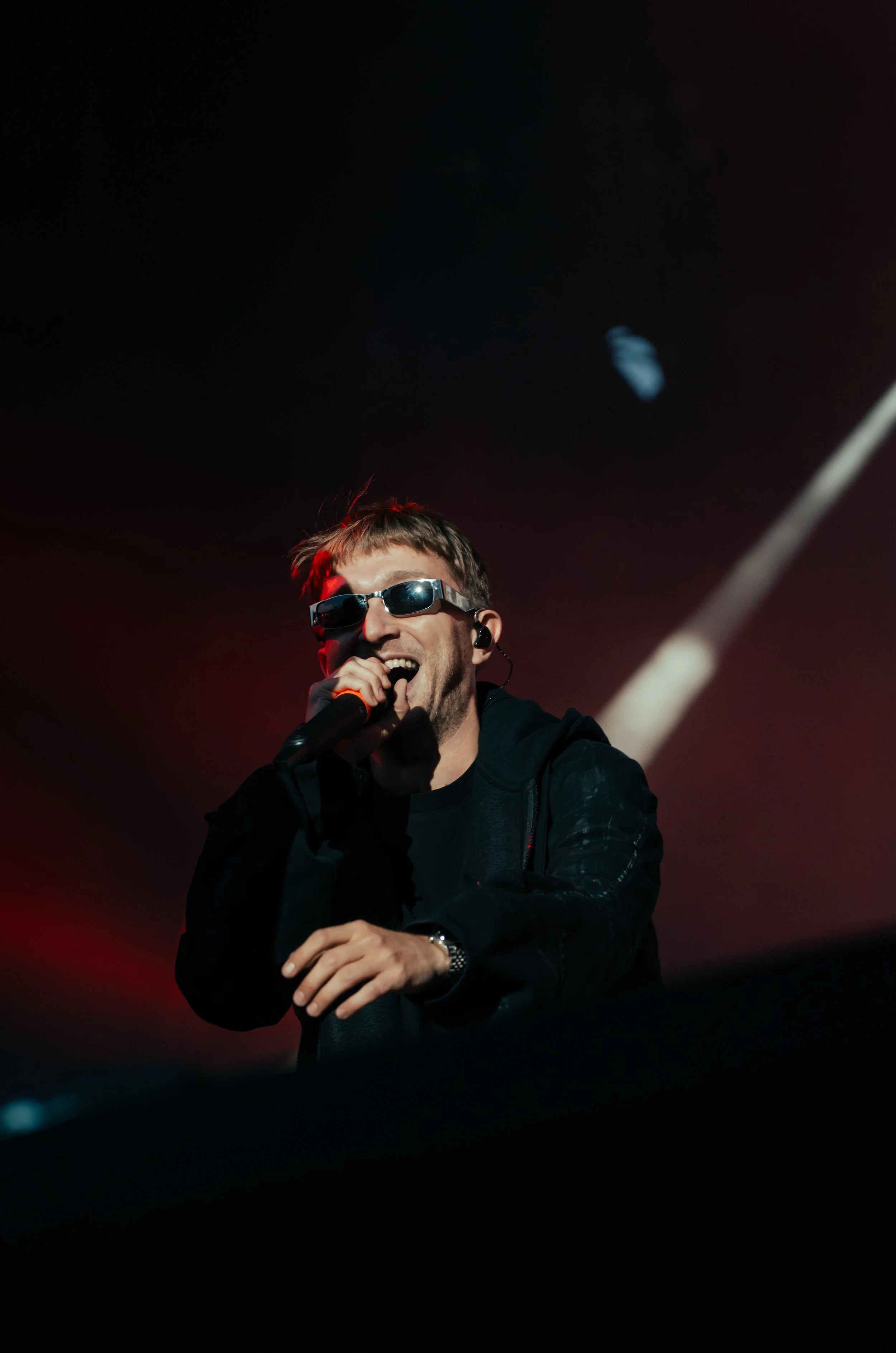 Artiste en concert portant des lunettes de soleil, tenant un microphone, souriant, avec un éclairage de scène dramatique