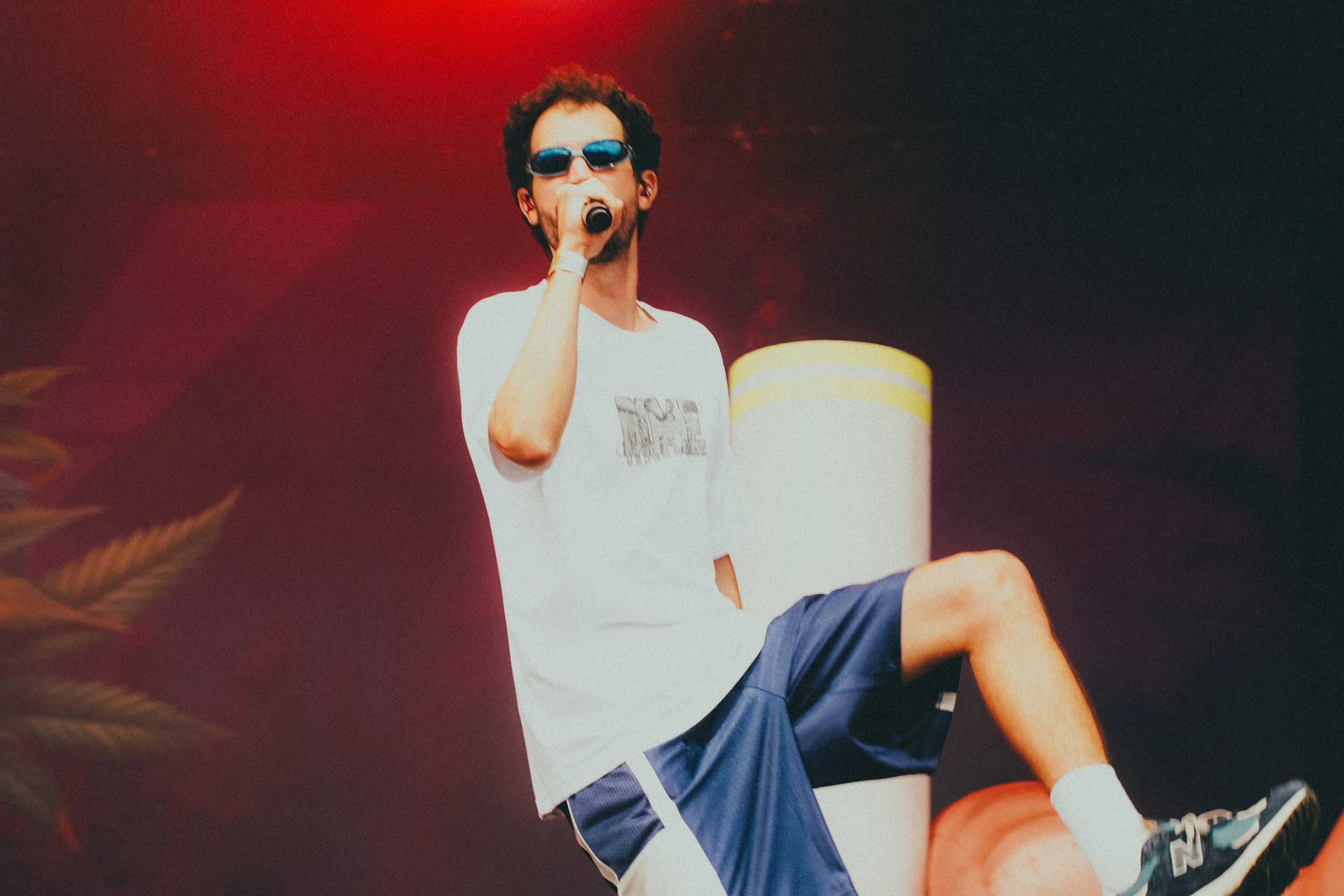 Un homme avec des lunettes de soleil, portant un t-shirt blanc et un short bleu, chante dans un microphone sur scène.
