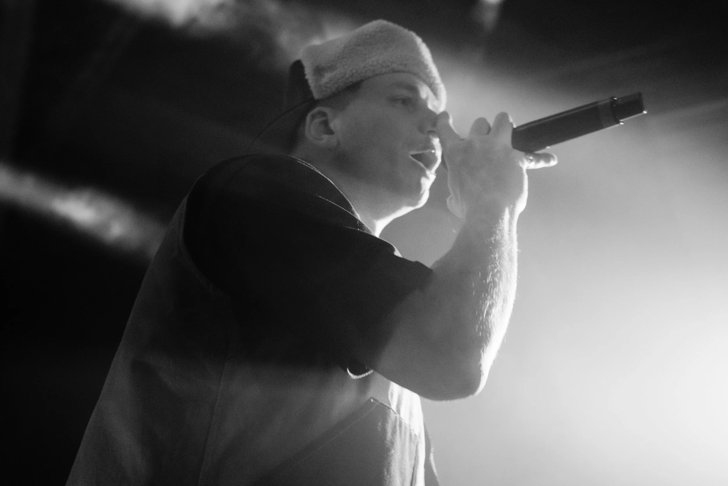 Un artiste de rap porte un bonnet et une veste, tenant un microphone en mouvement lors d'une performance sur scène, avec un éclairage fort et une ambiance sombre.