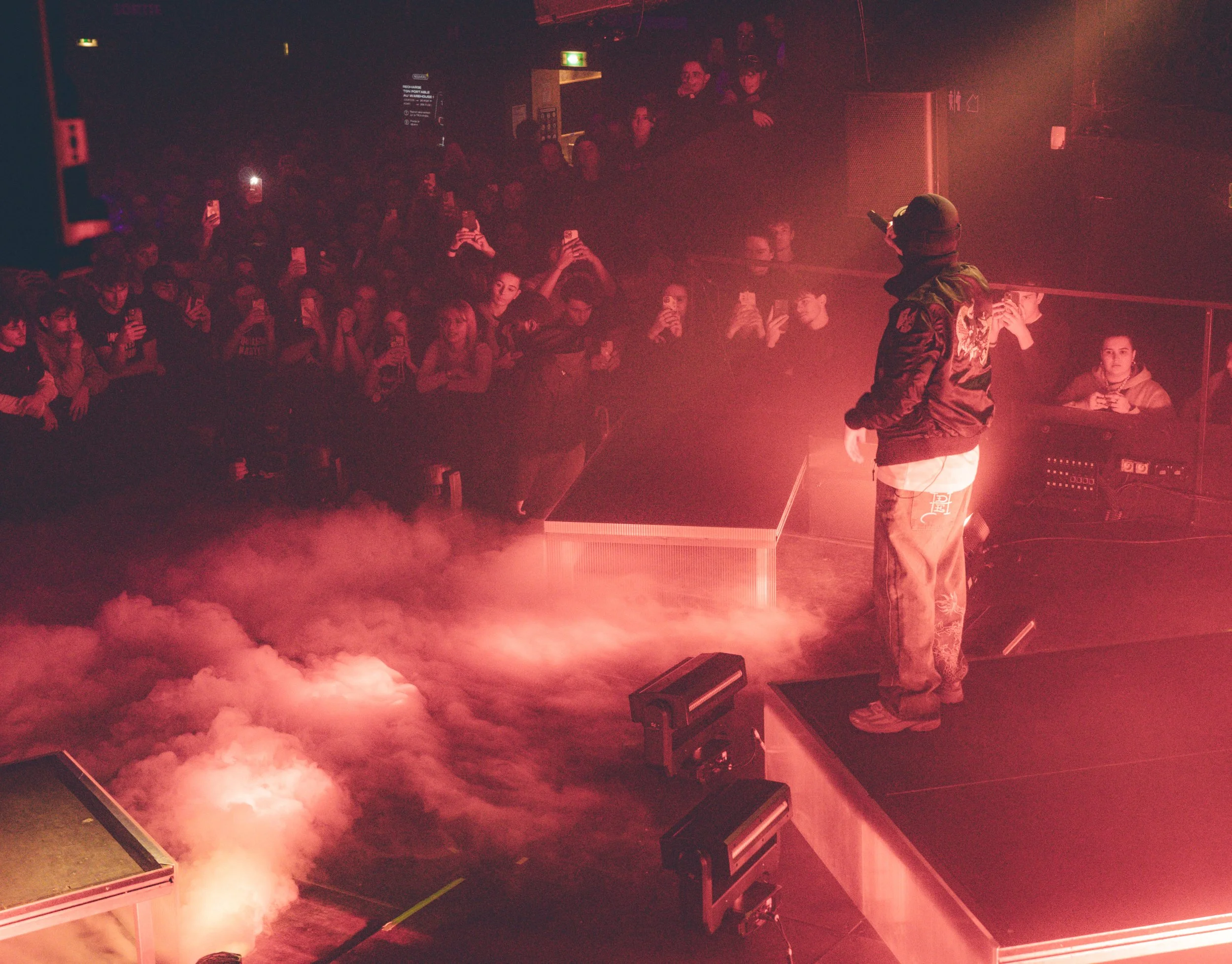 Un artiste sur scène lors d’un concert, entouré de fumée, avec un public regardant et prenant des photos, éclairage rougeâtre.