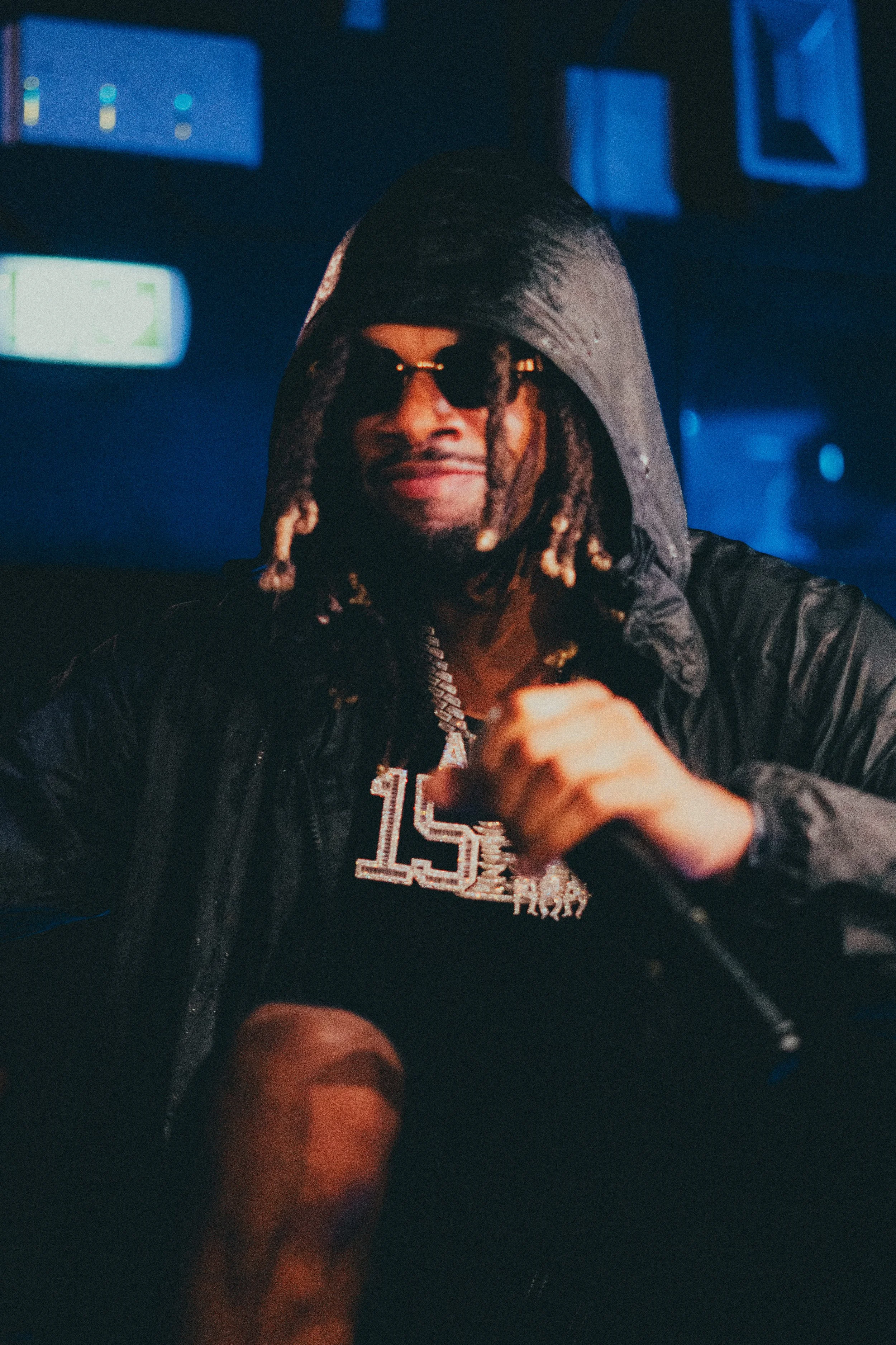 Un homme portant des lunettes de soleil, un collier avec la lettre "L" et une veste noire, tenant un micro dans une ambiance sombre et bleutée.