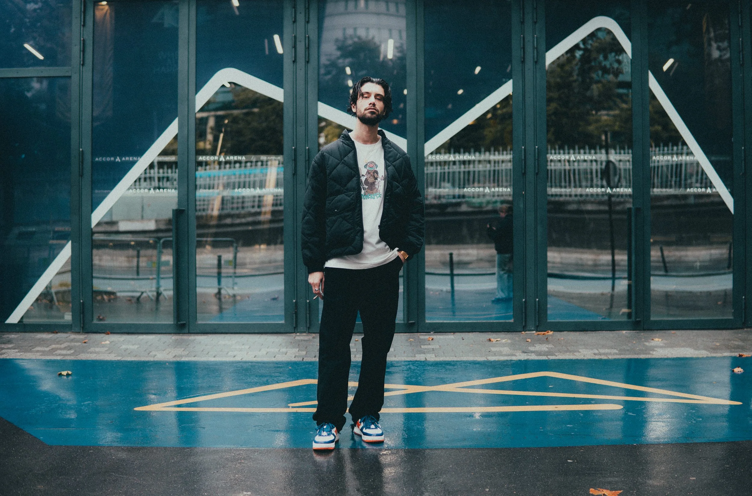 Un homme debout devant une façade en verre avec des dessins blancs, portant un pantalon noir, un sweat-shirt blanc avec un motif et une veste noire, les mains dans les poches.