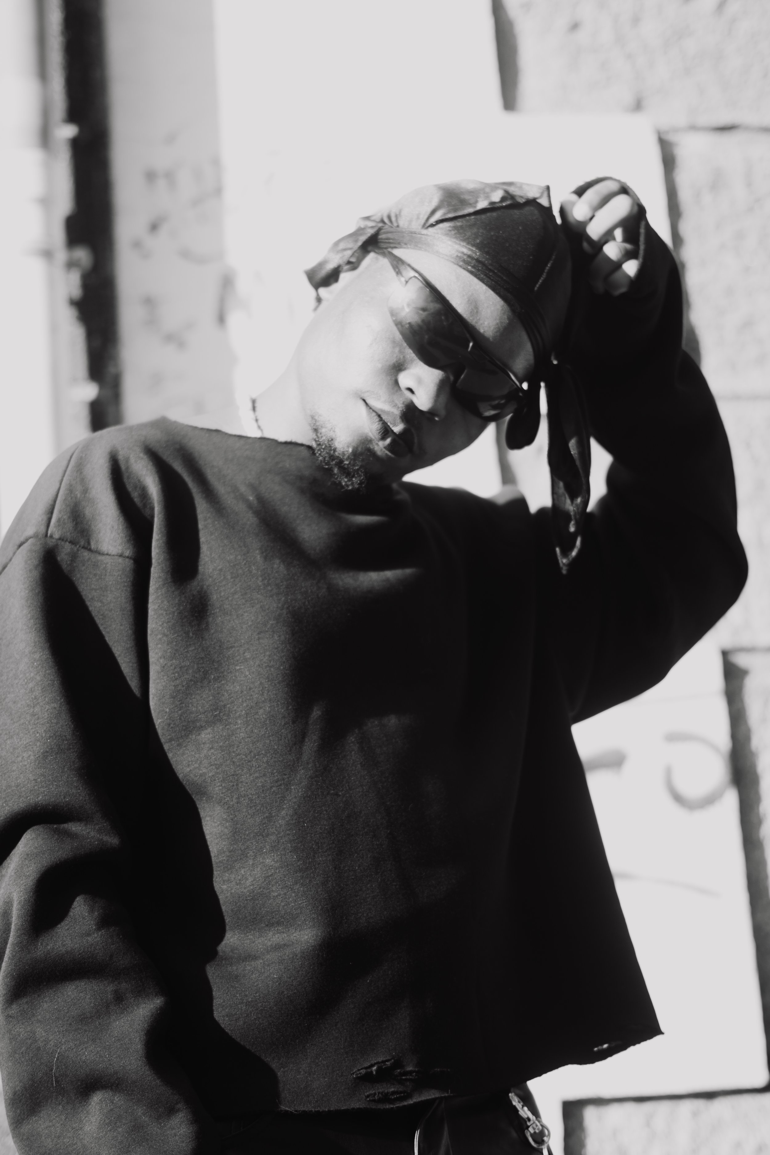 Jeune homme portant un bonnet, des lunettes de soleil, et un sweat noir, se tenant contre un mur en pierre, dans une pose décontractée.