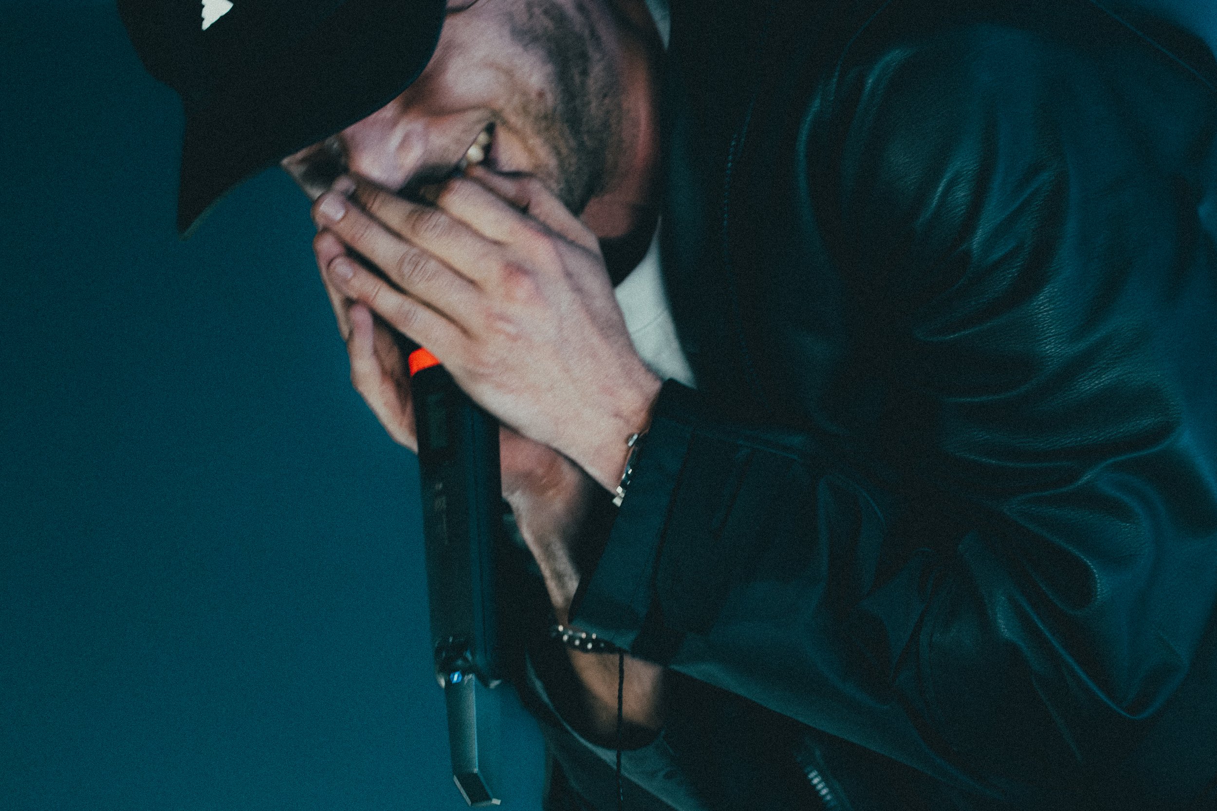 Un homme portant un chapeau noir et un blouson en cuir noir, tenant un pistolet, semble en train de simuler un tir ou de faire une blague, avec une expression faciale concentrée ou amusée.