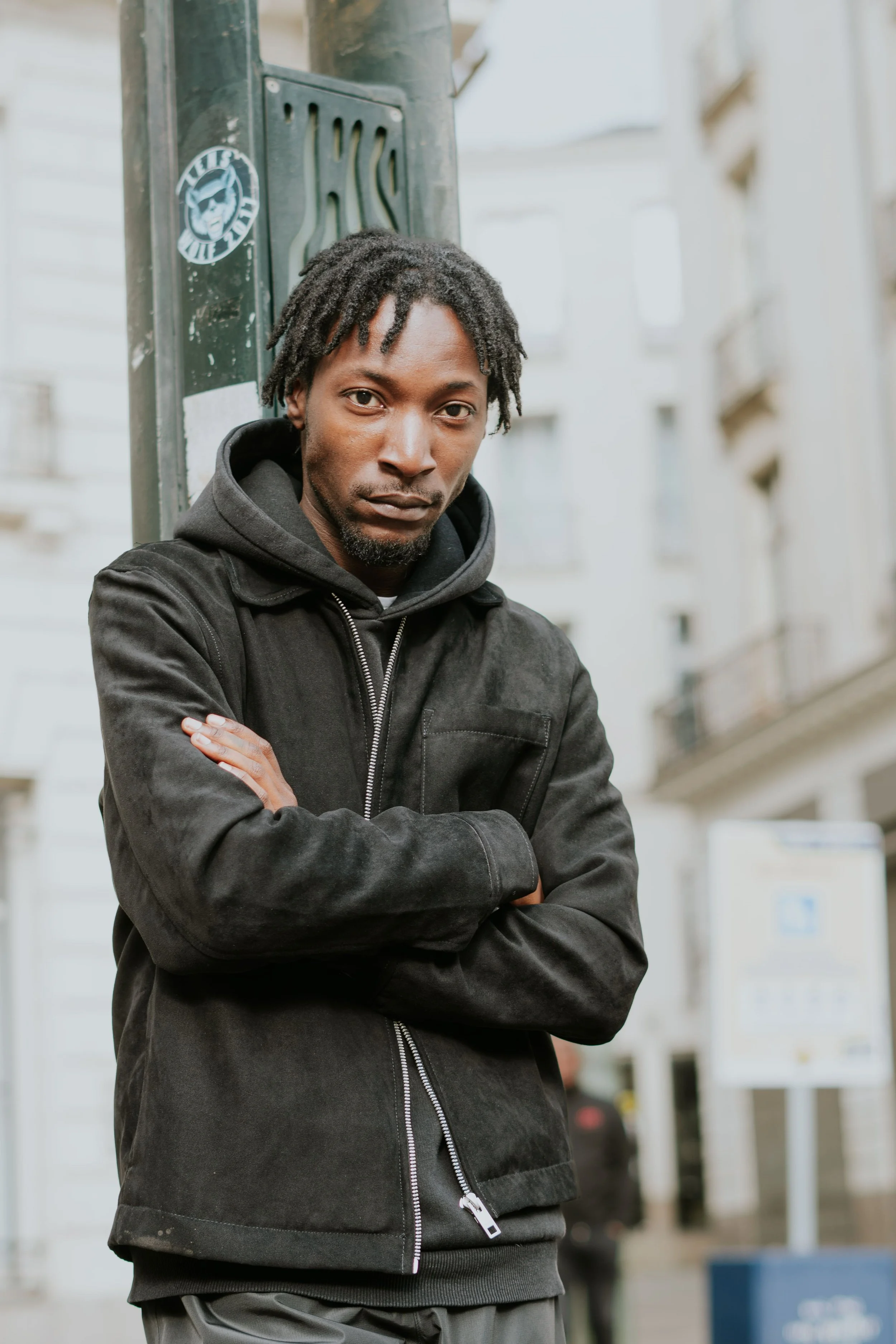 Un homme avec des cheveux bouclés et une barbe, portant une veste noire et une capuche, se tient avec les bras croisés dans une rue urbaine.
