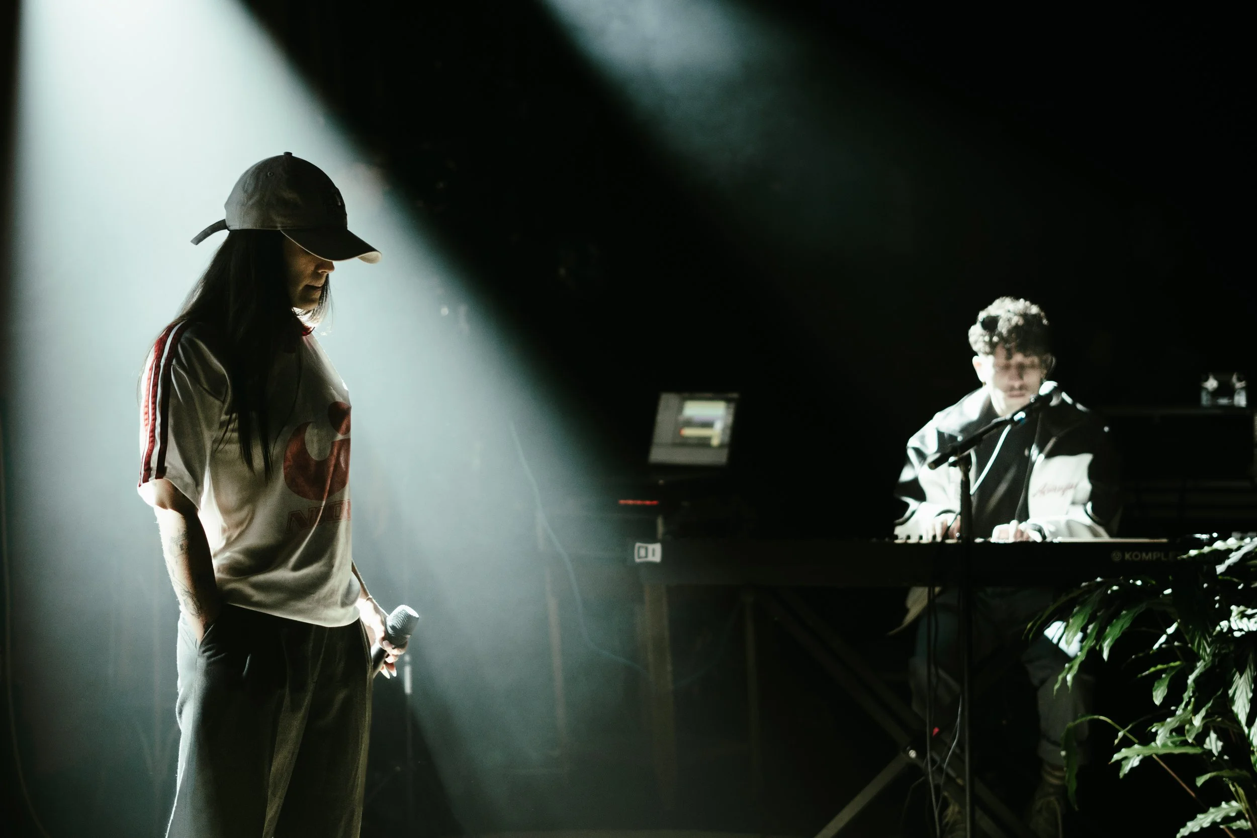 Une femme avec une casquette et une chemise à manches courtes, tenant un microphone, se tient dans un éclairage tamisé, avec un musicien jouant du clavier en arrière-plan.