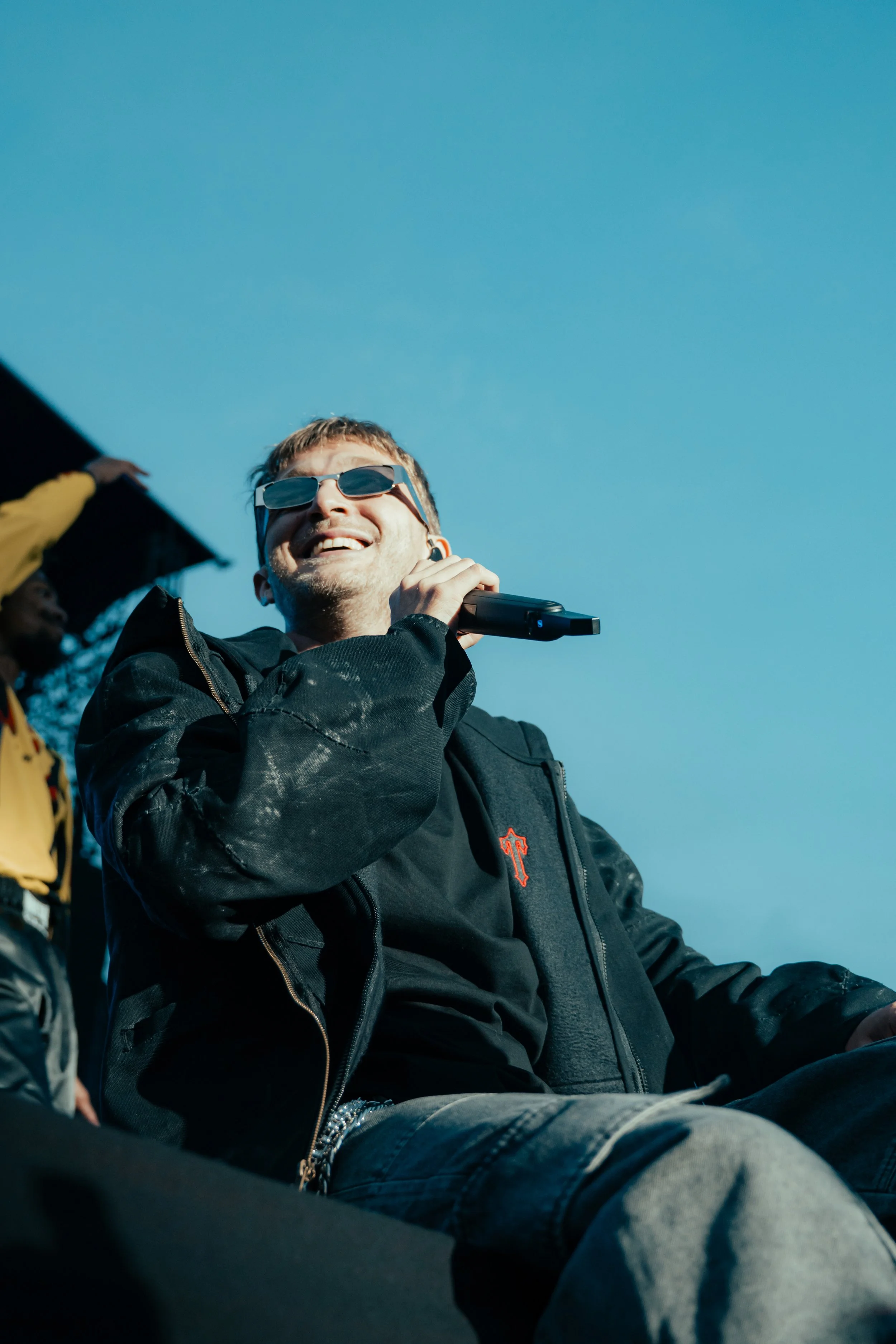 Un homme souriant, portant des lunettes de soleil et tenant une microphone, assis à l'extérieur avec un ciel bleu en arrière-plan.